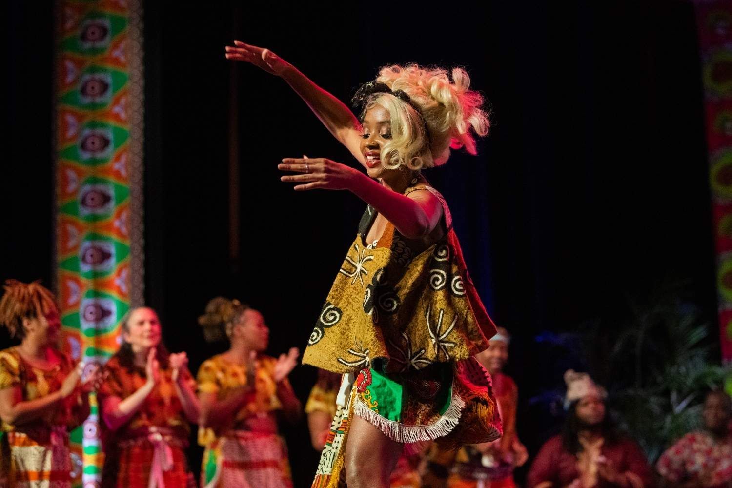 Woman in African-style costume dances on stage, arm raised. Others applaud.