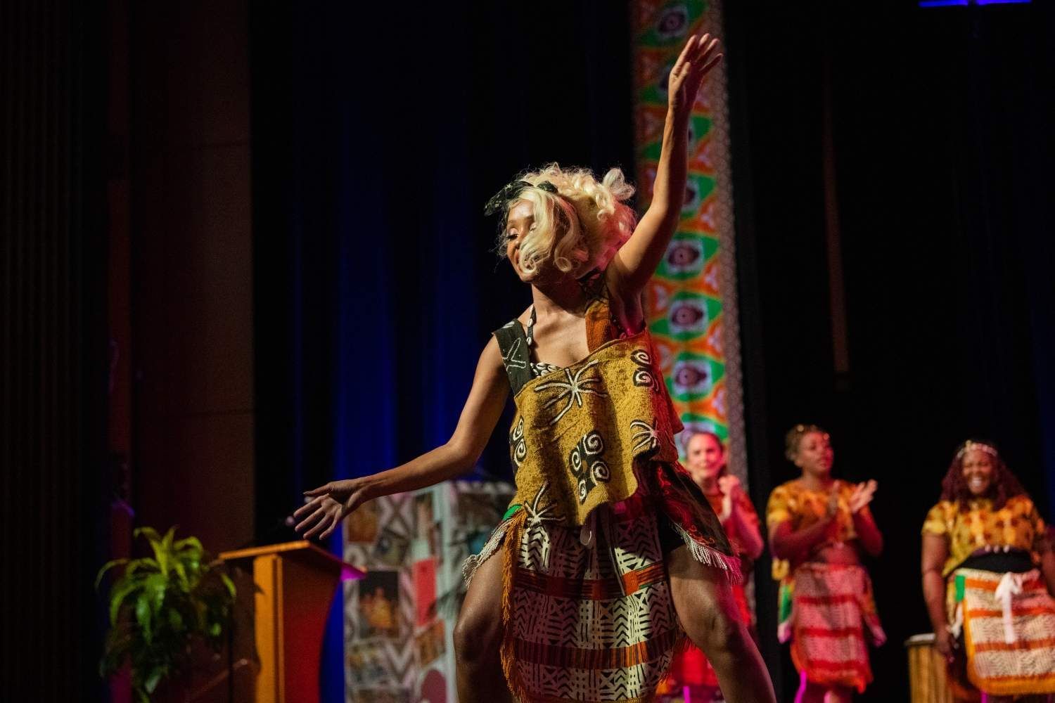 Woman in African print dress dances on stage; others in background clap.