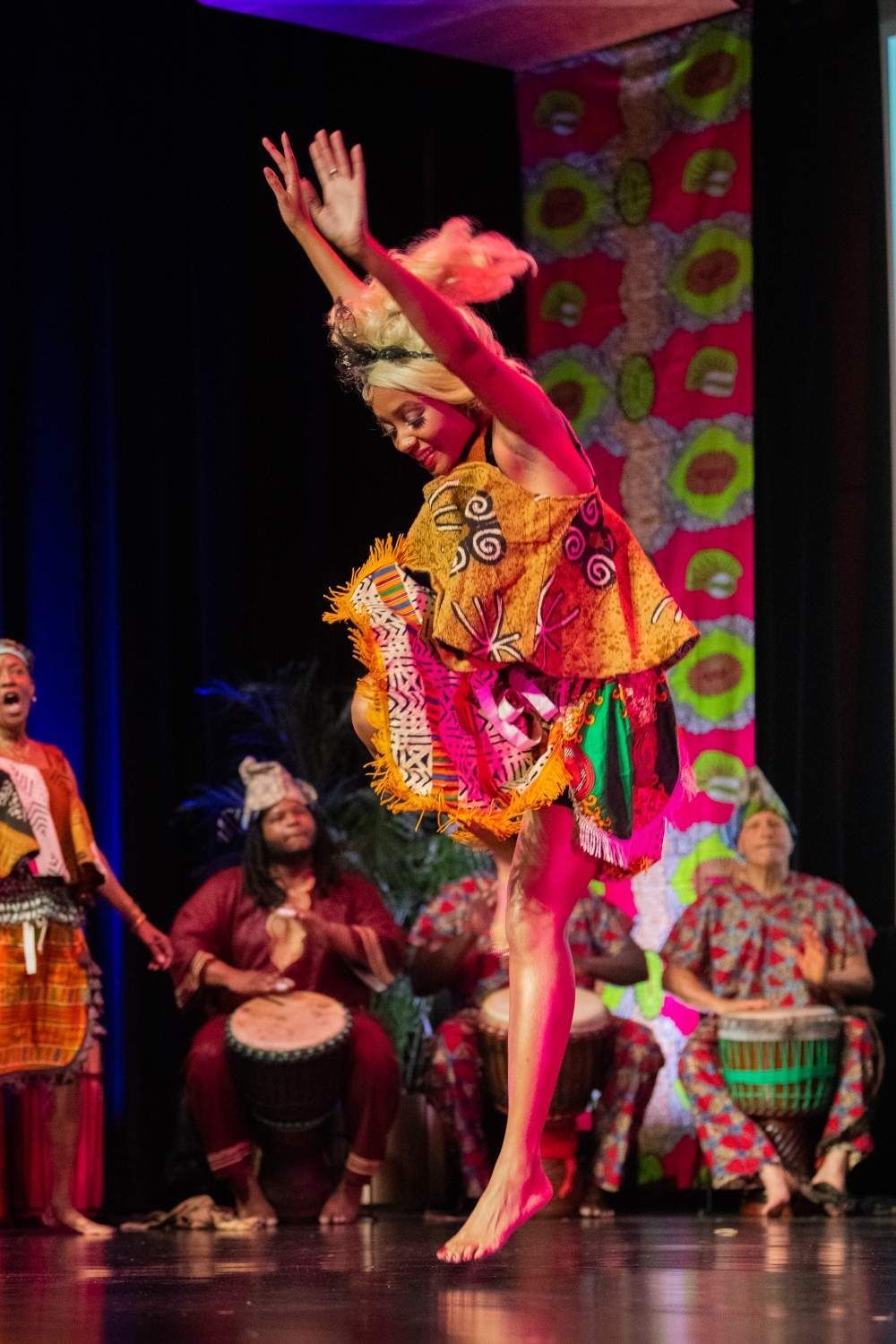 Woman in colorful African attire leaps while dancing on stage, band playing drums in background.