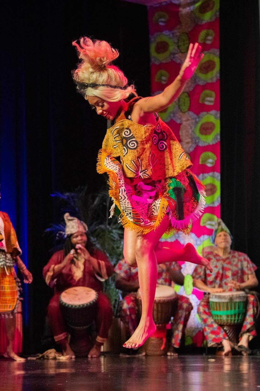 Woman in vibrant African dance costume leaps on stage, musicians with drums in background.