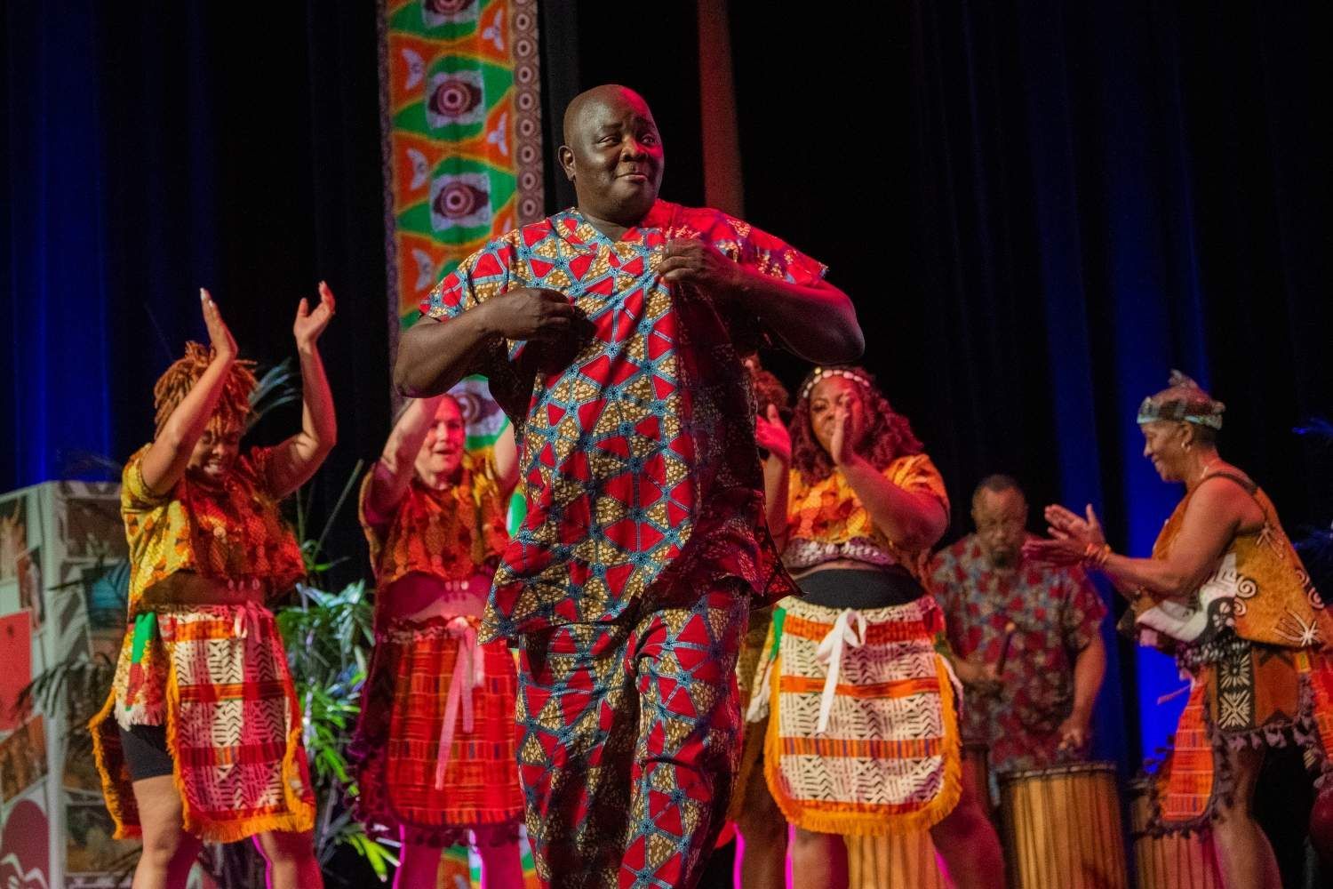 A man in colorful African attire dances on stage with a group; they clap and celebrate.