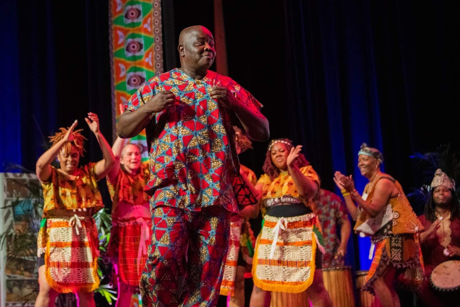 Man in patterned red outfit dances onstage with a group of dancers in colorful costumes.
