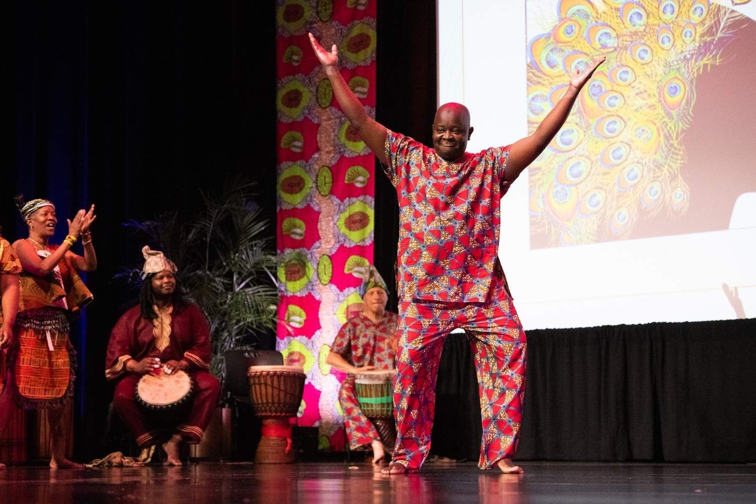 Man in red patterned outfit dances on stage, arms raised. Others play drums in front of a colorful backdrop.