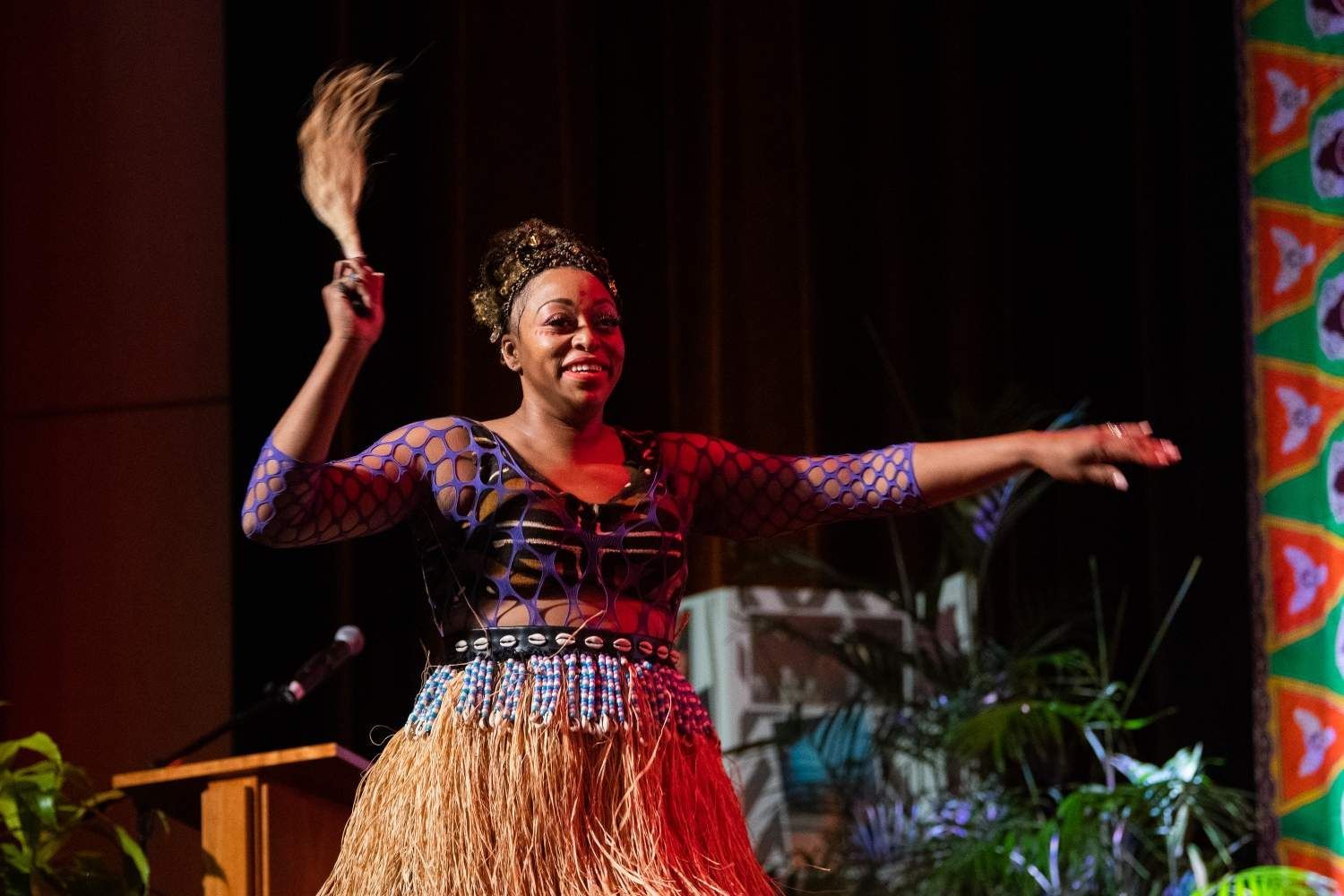 Woman in traditional African attire dances onstage, holding a fan, smiling, stage background.