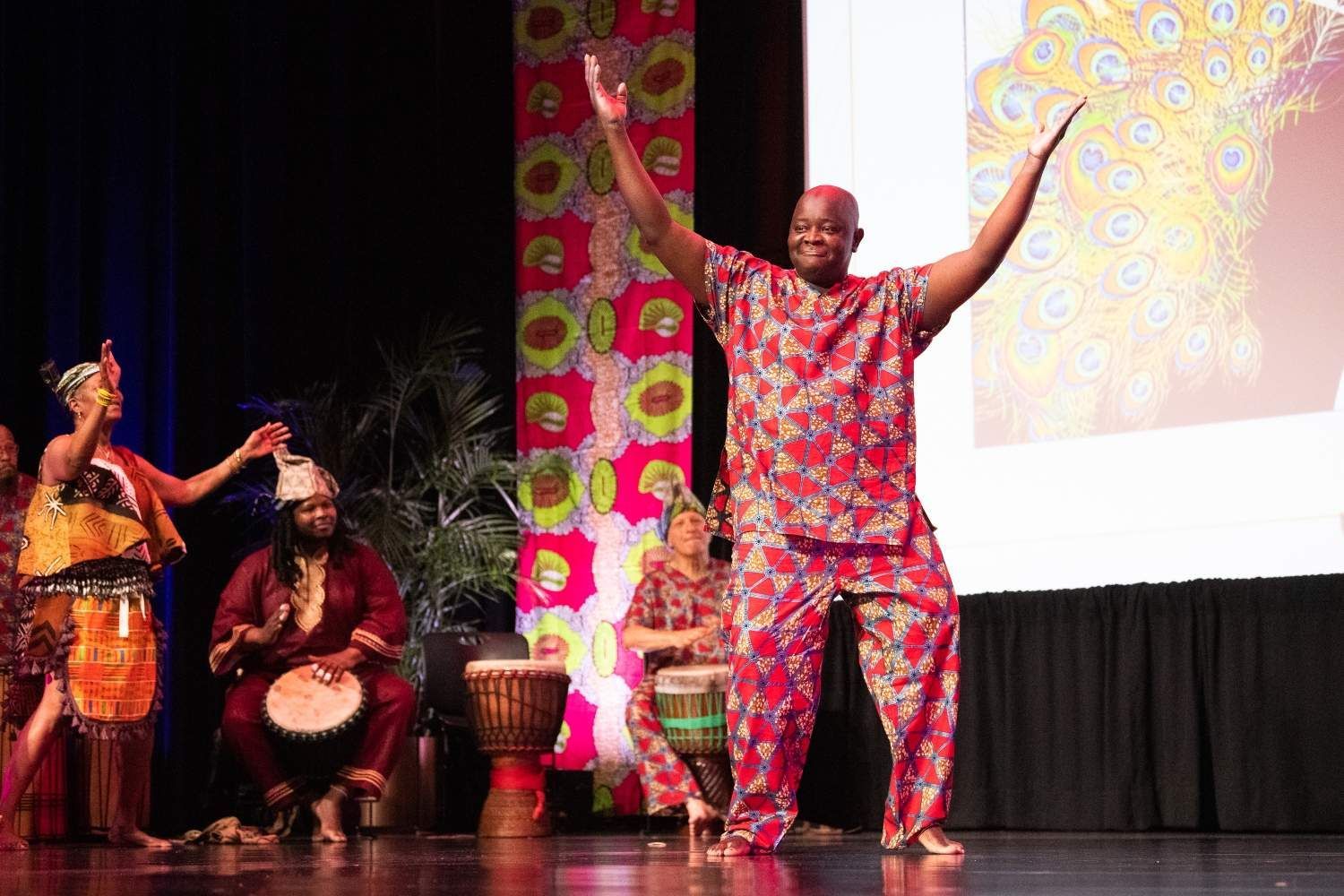 Man in red African attire dances on stage, arms raised; musicians play drums.