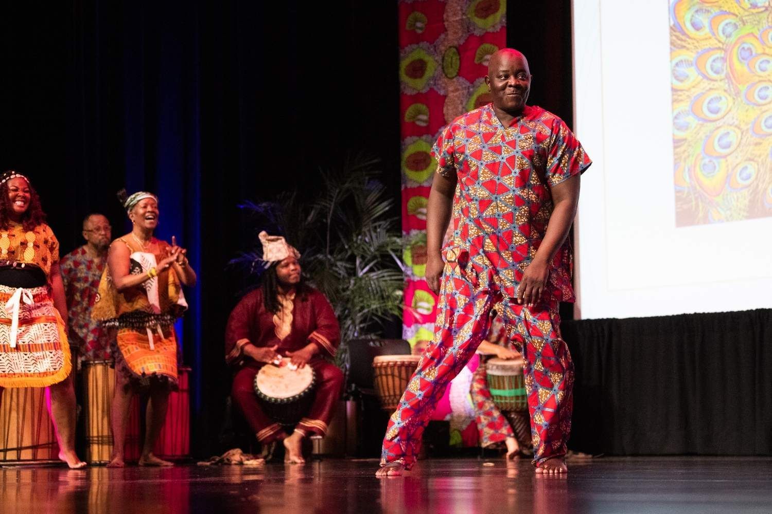 People on stage in African attire perform, using drums, in front of a patterned backdrop.