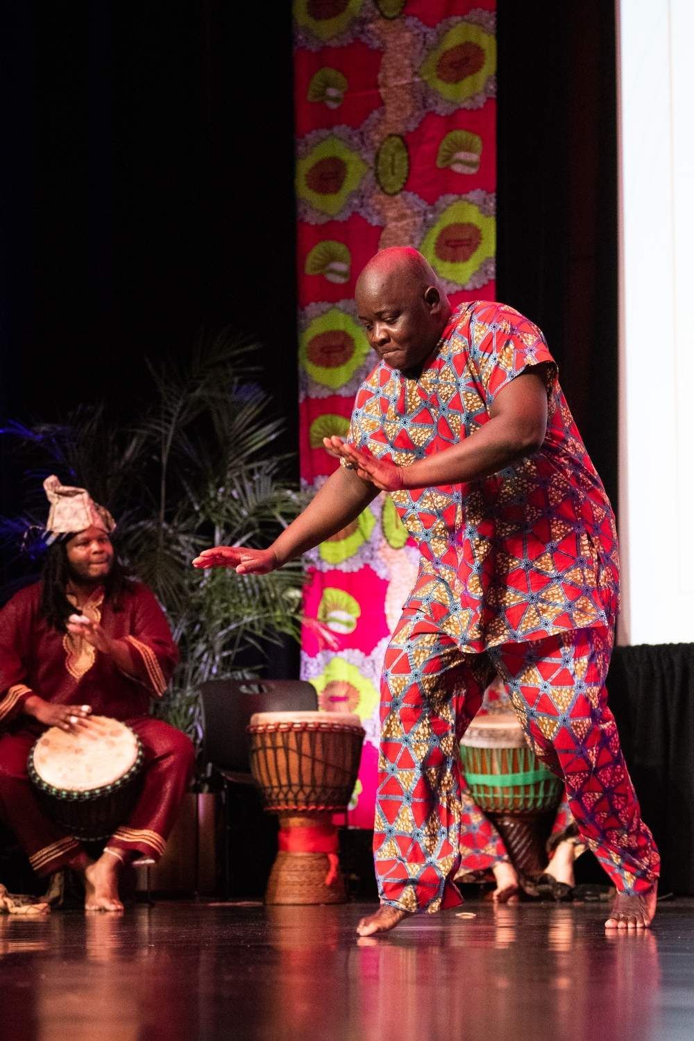 Man in red patterned outfit dances on stage, two others play drums.
