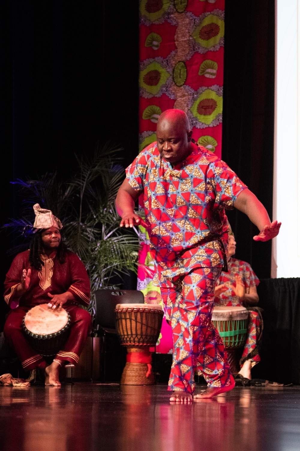 Man dancing in colorful African attire, drummers in background, red stage, patterned backdrop.