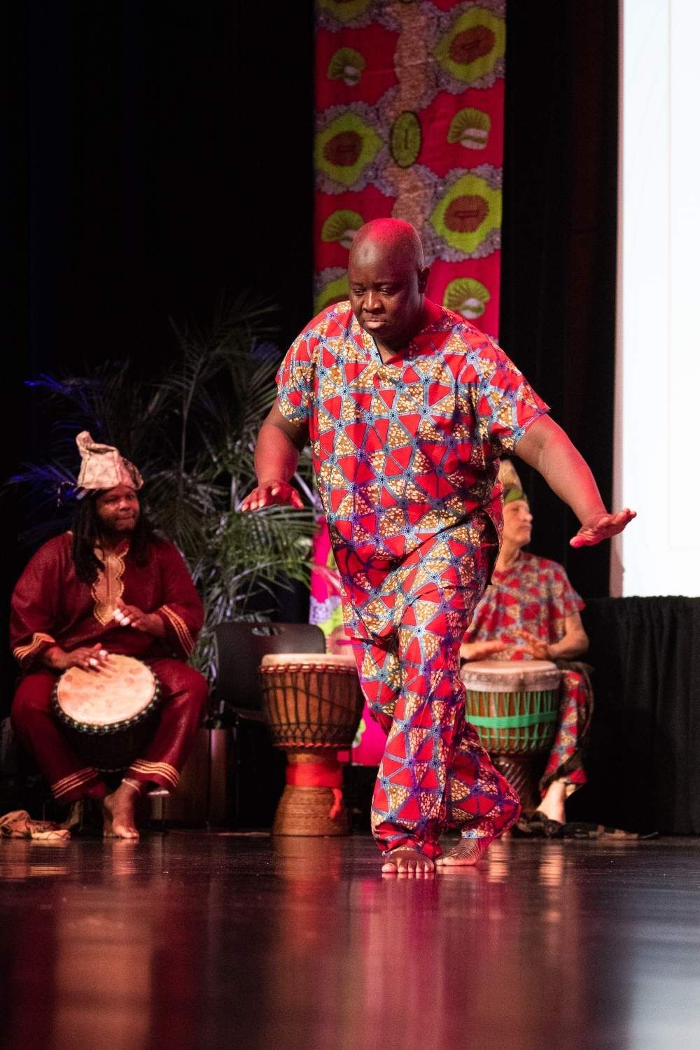 Man dancing in colorful African attire, performing in front of drummers on stage.