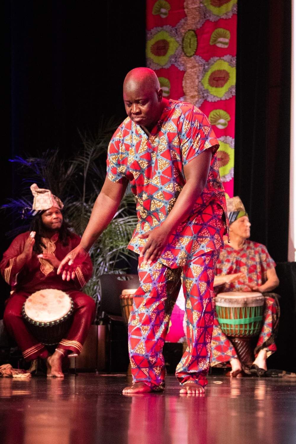 Man in patterned red outfit dances onstage, musicians playing drums.