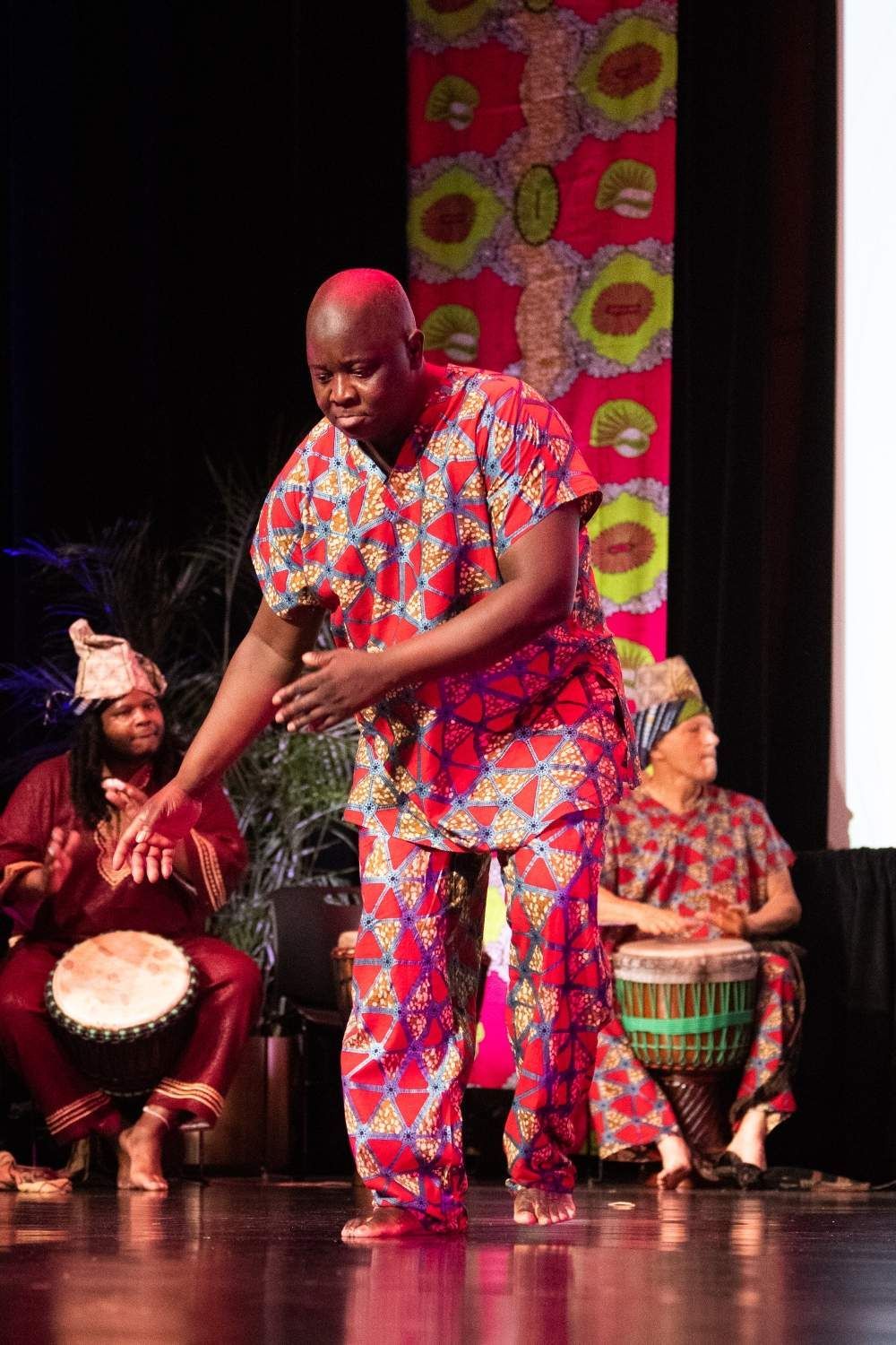 Man in patterned African clothing dances on stage, accompanied by drummers.