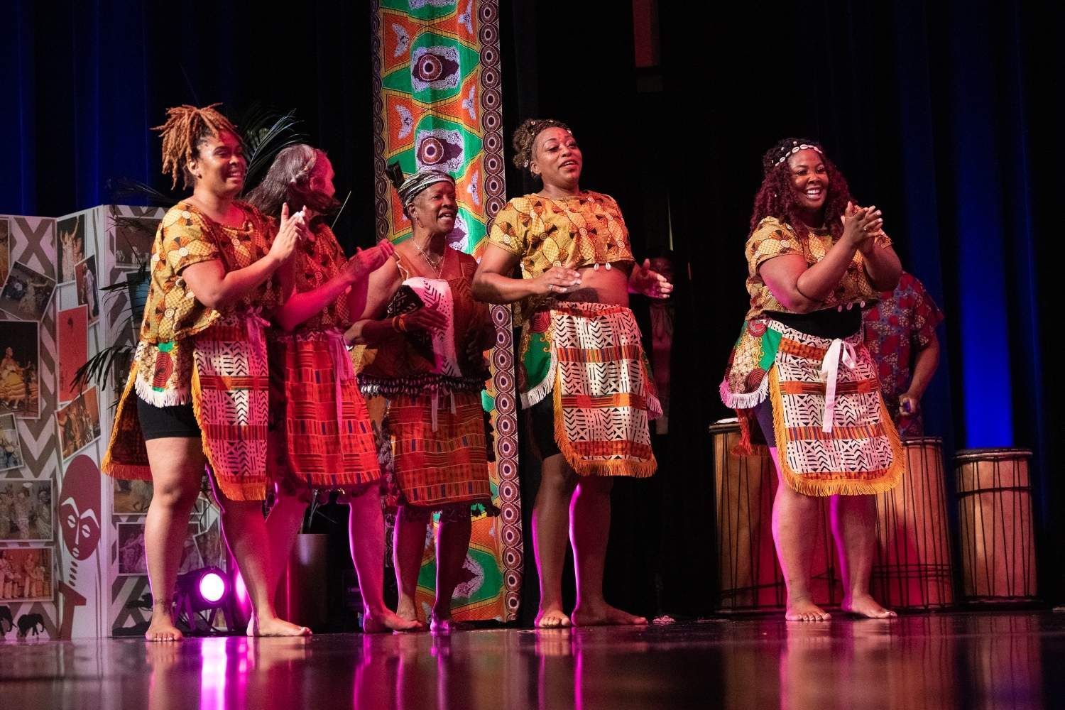 Five women in colorful African attire on stage, clapping and smiling. Drums in background.
