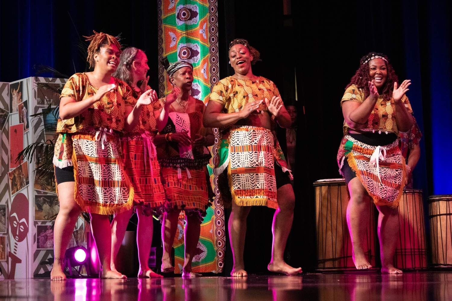 Group of women in colorful African attire performing on stage, clapping and smiling.