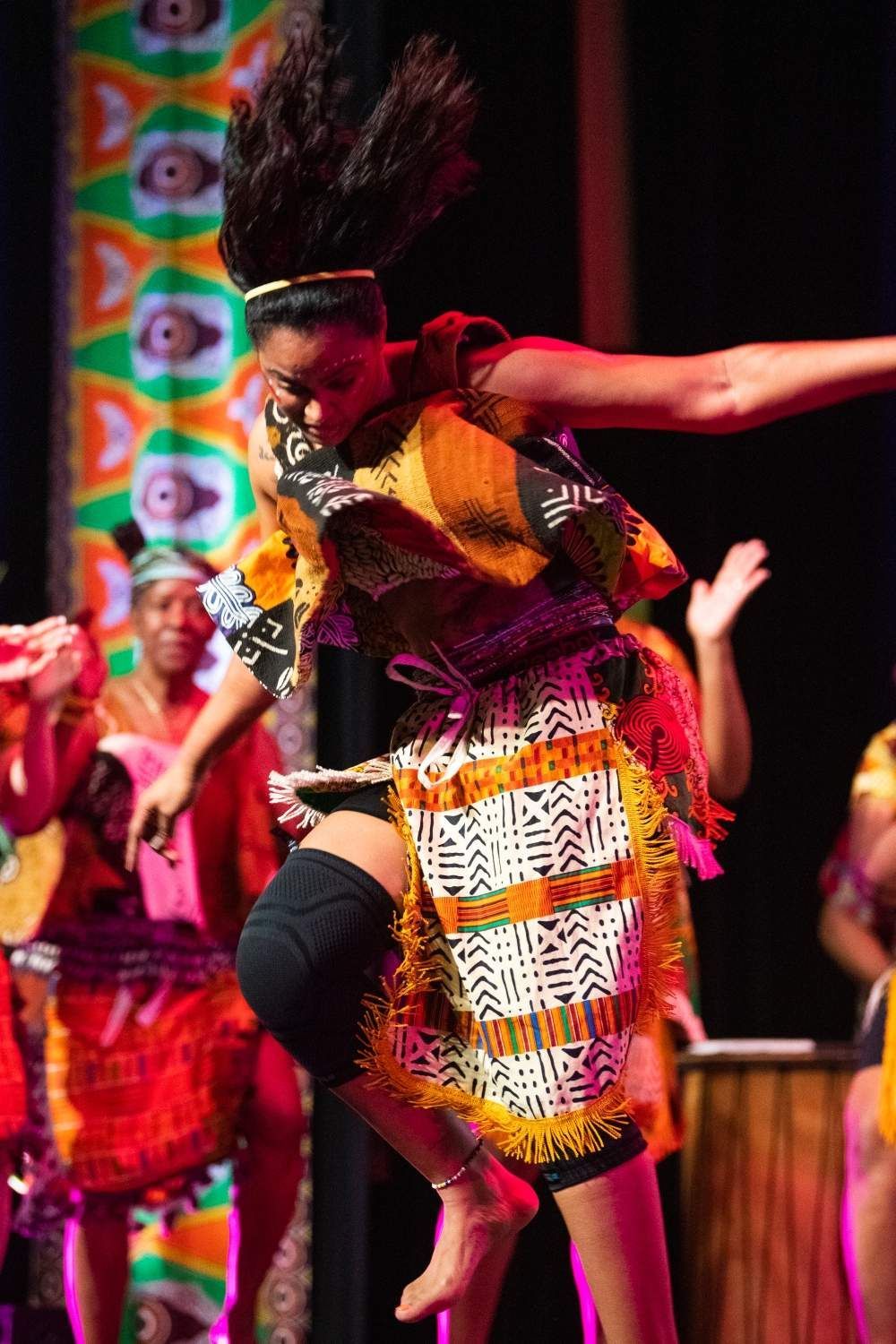 Woman in African print costume leaps during a dance performance; other performers and stage backdrop visible.
