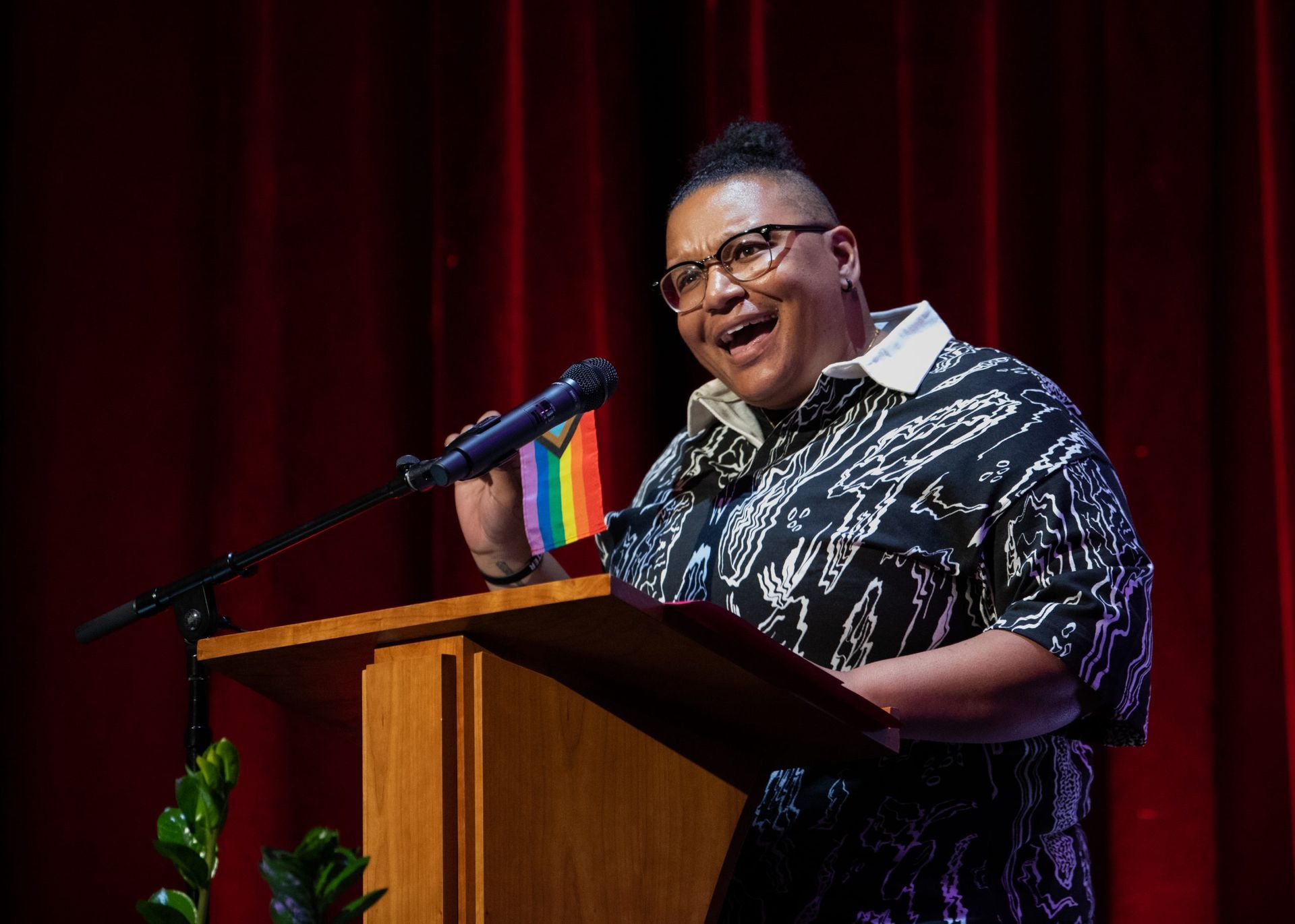 Person speaking at a podium, holding a rainbow flag. They wear patterned shirt, glasses, smile. Red curtains in background.