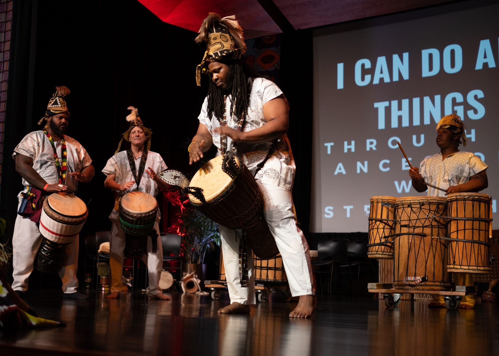 African drummers on stage, wearing white outfits and performing, with a projected message: 