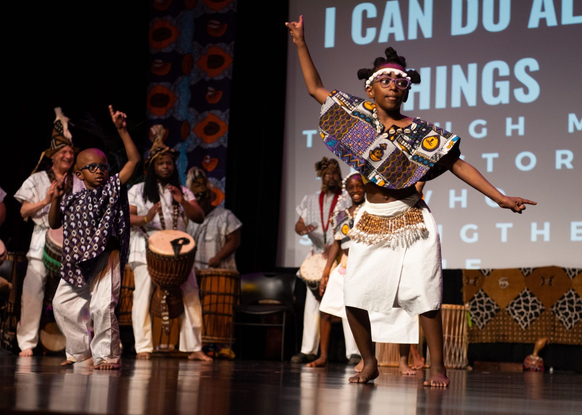 African dancers on stage with drummers. A woman dances in white skirt and patterned top, arms raised.