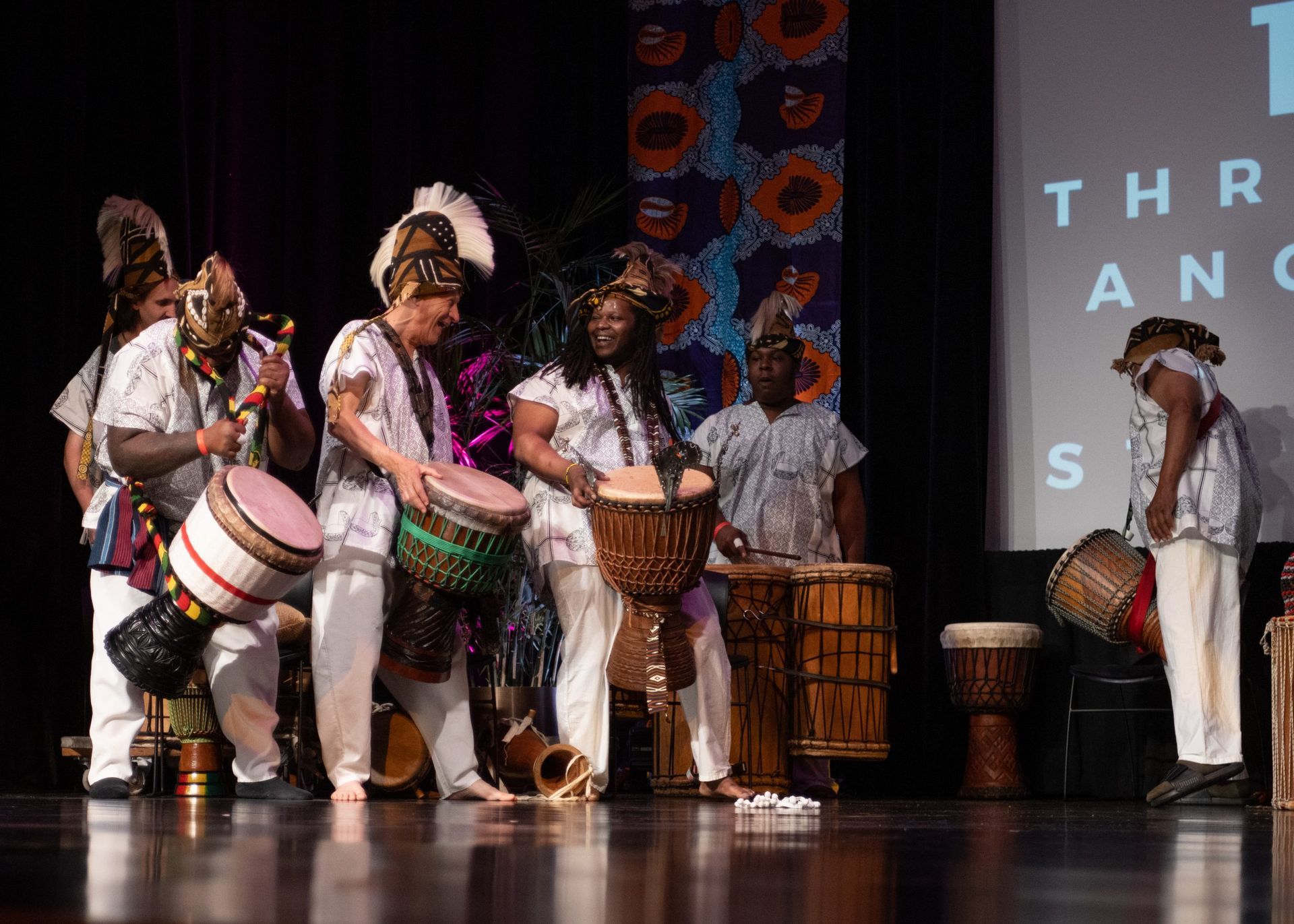 African drummers in white outfits with drums on stage. Backdrop with pattern and text.