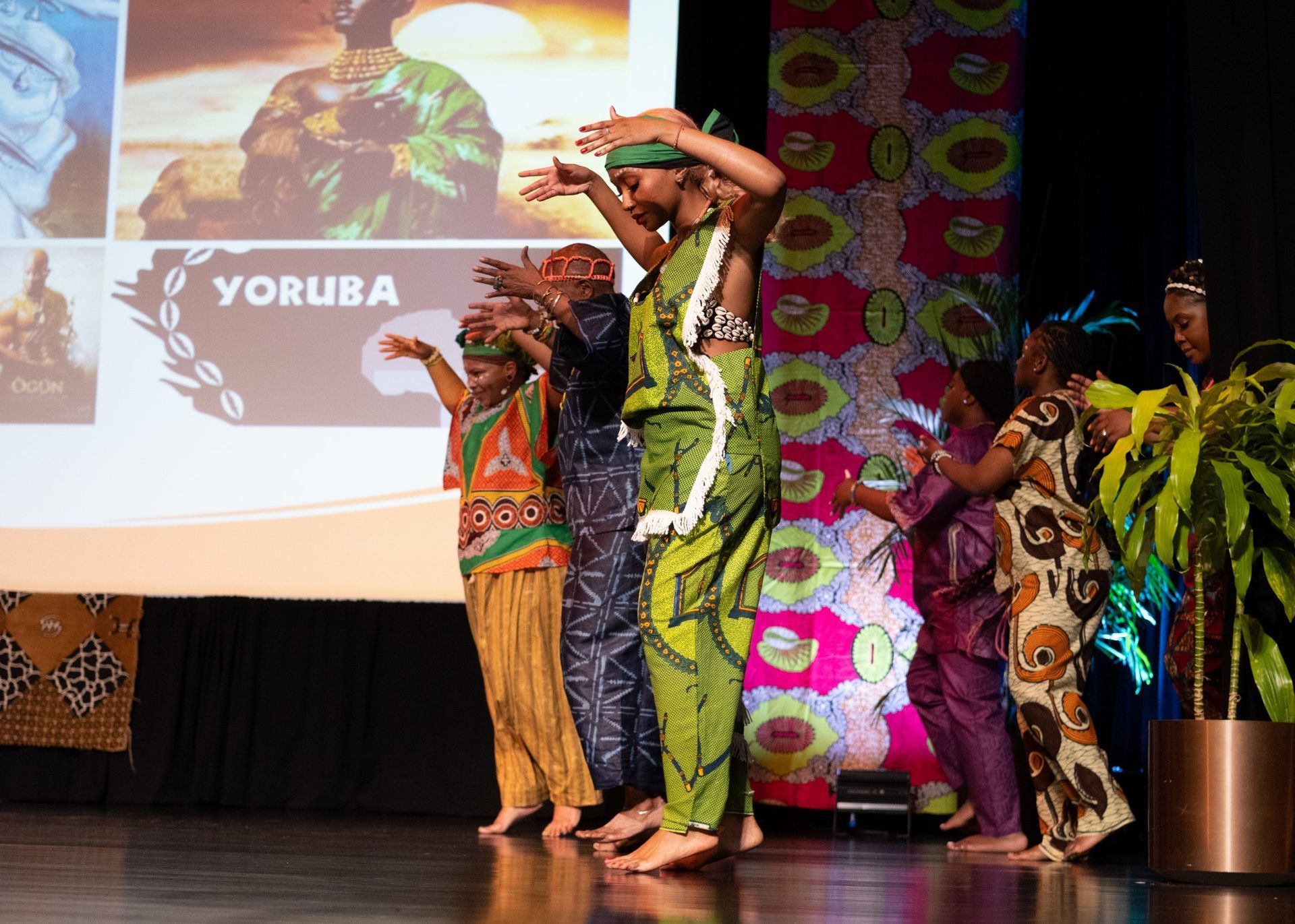 People in traditional Yoruba attire dance on a stage, arms raised, in front of a screen with images and text.
