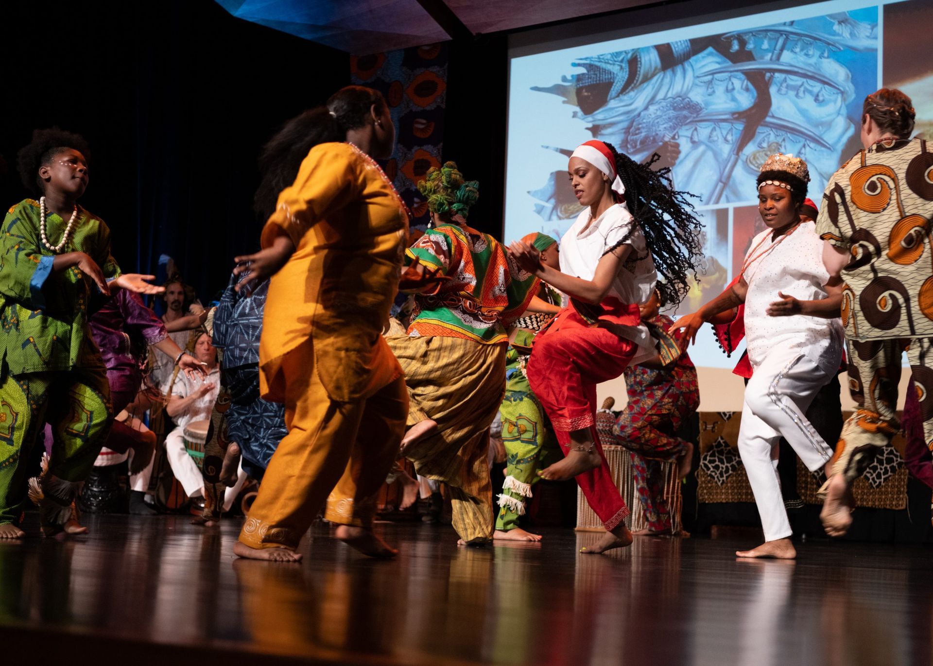 A group of dancers in colorful costumes perform on stage; a screen displays an image behind them.
