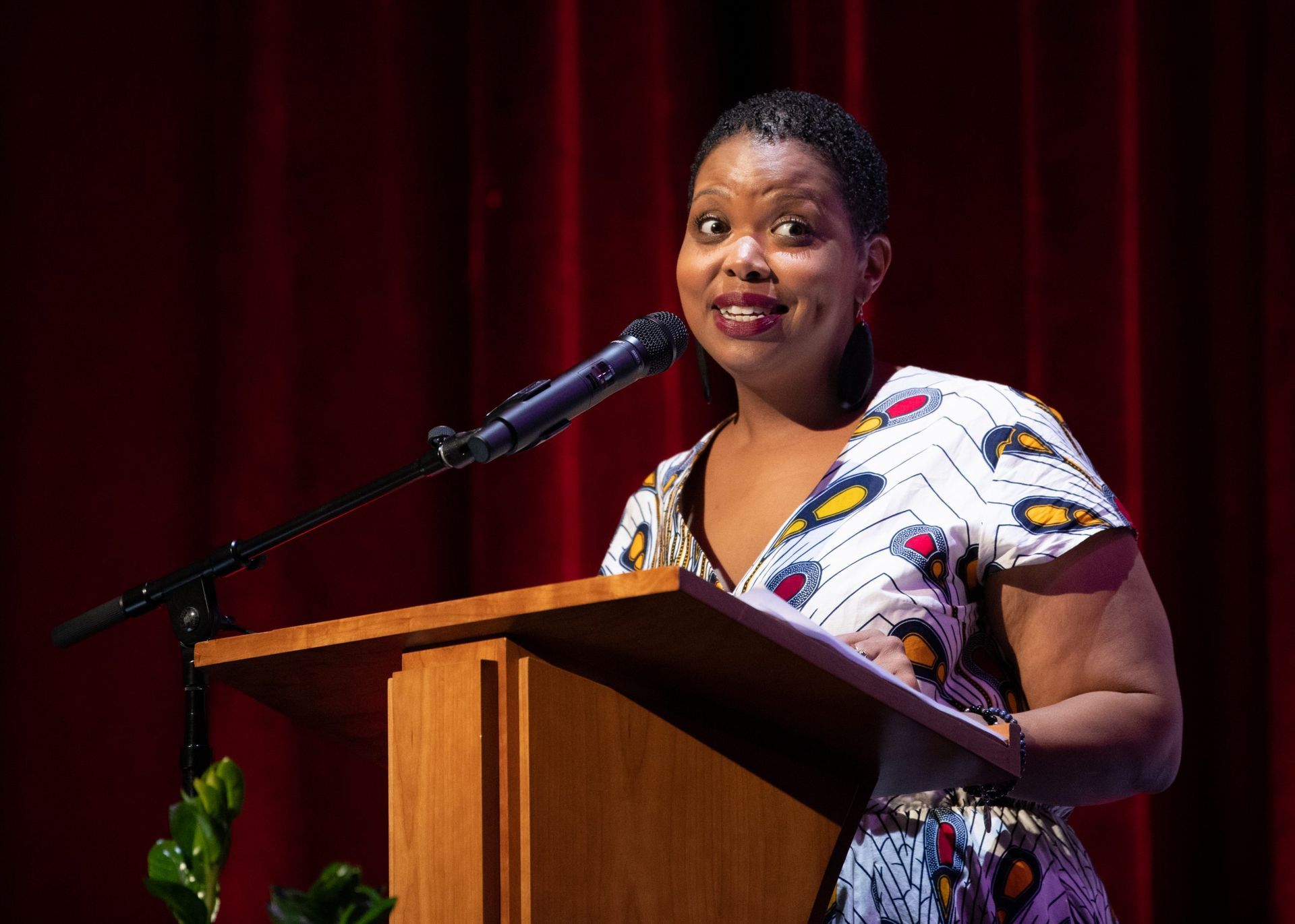 Woman speaking at a podium, wearing patterned dress; dark background, red curtains.