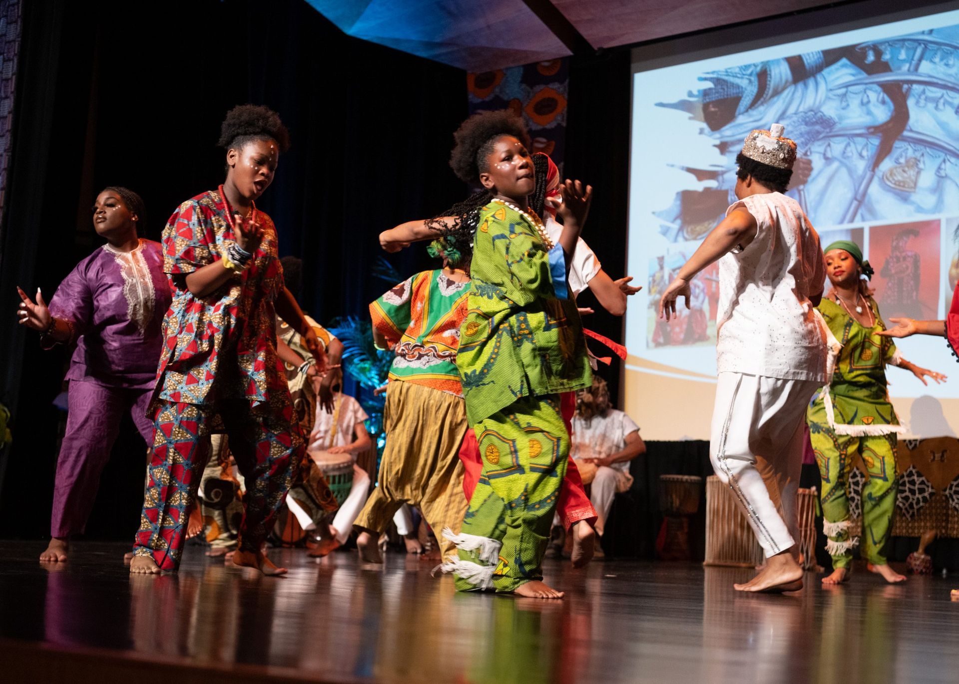 People in colorful African attire dance on a stage in front of a projected image.