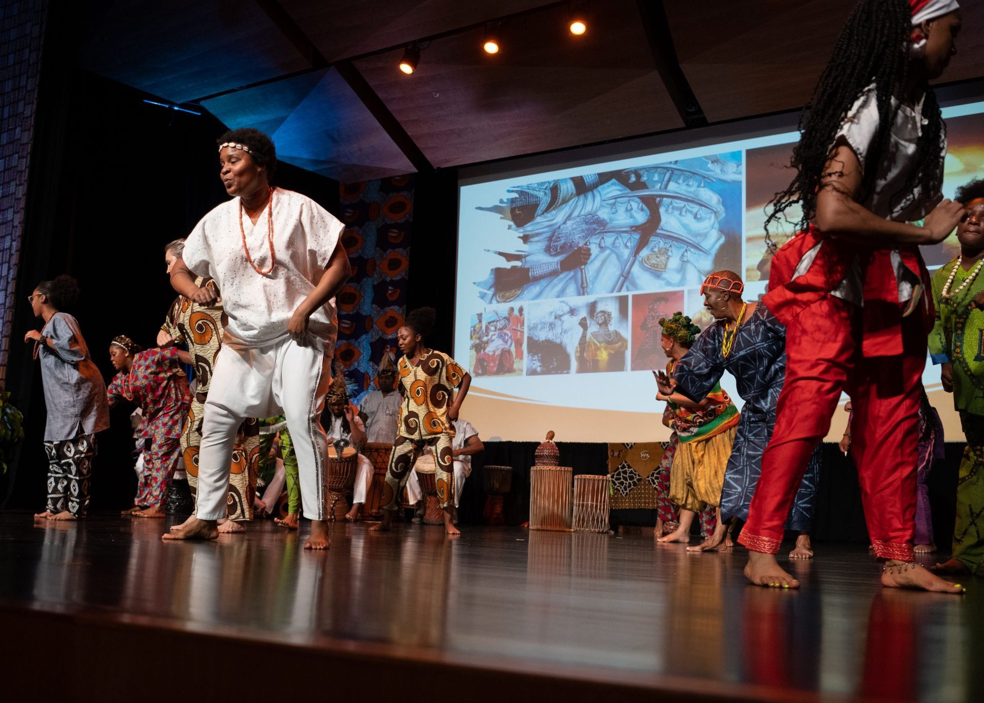 Group of people dancing on a stage.  They wear colorful clothing, with a screen displaying photos in the background.