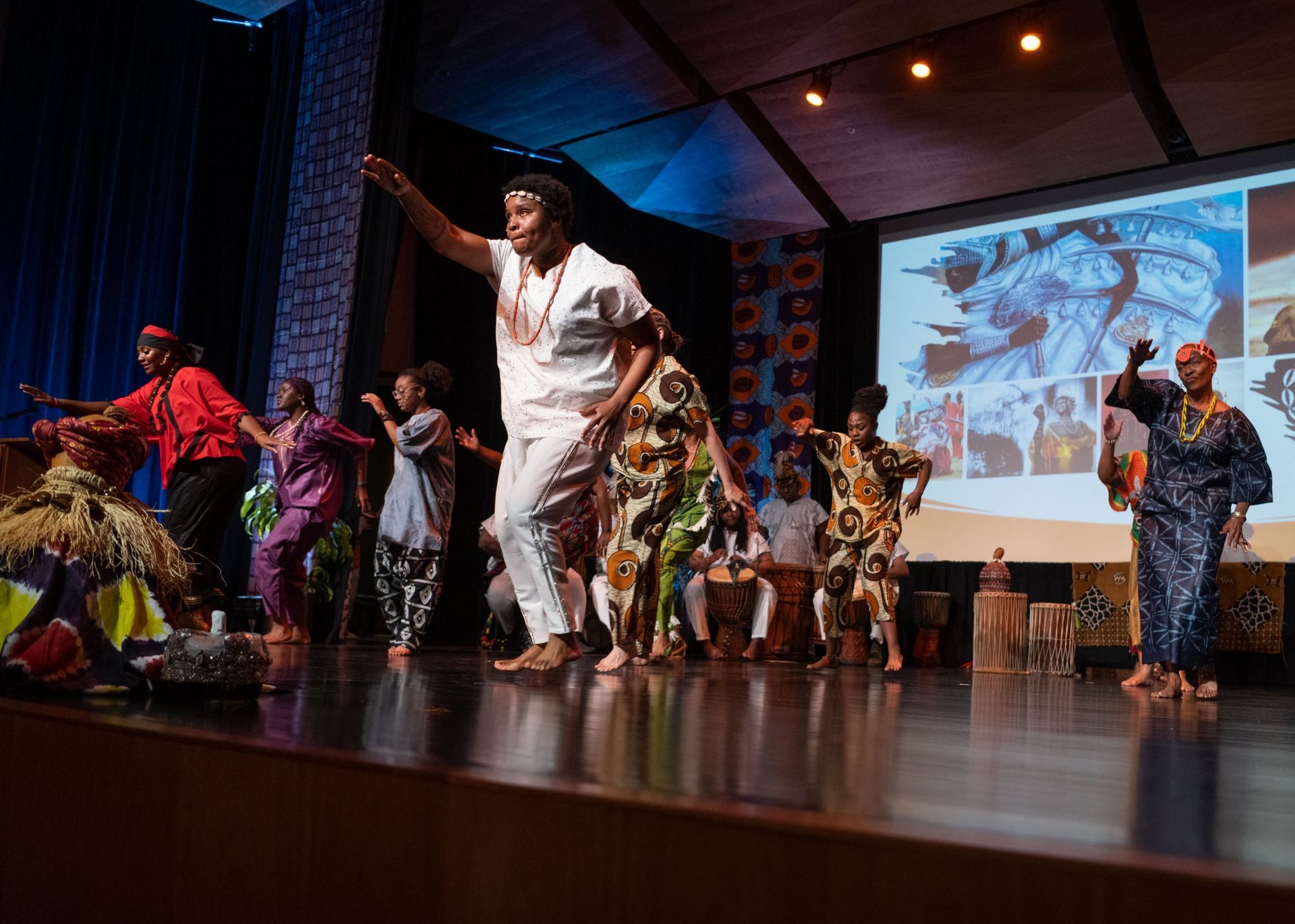 African dancers in colorful costumes perform on a stage with a screen displaying images.