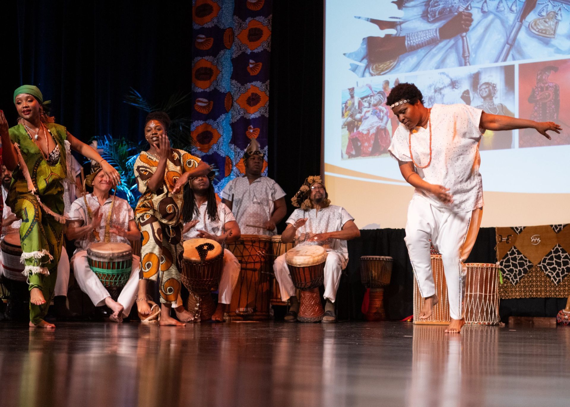 African dancers perform on stage with drummers; colorful attire, joyful expressions.