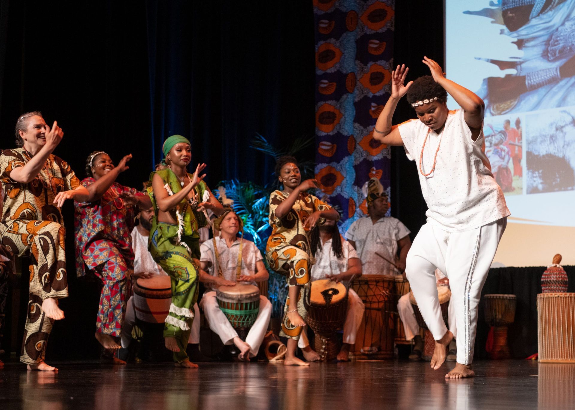 People dance on stage; a dancer in white performs energetically while others play drums and sing.