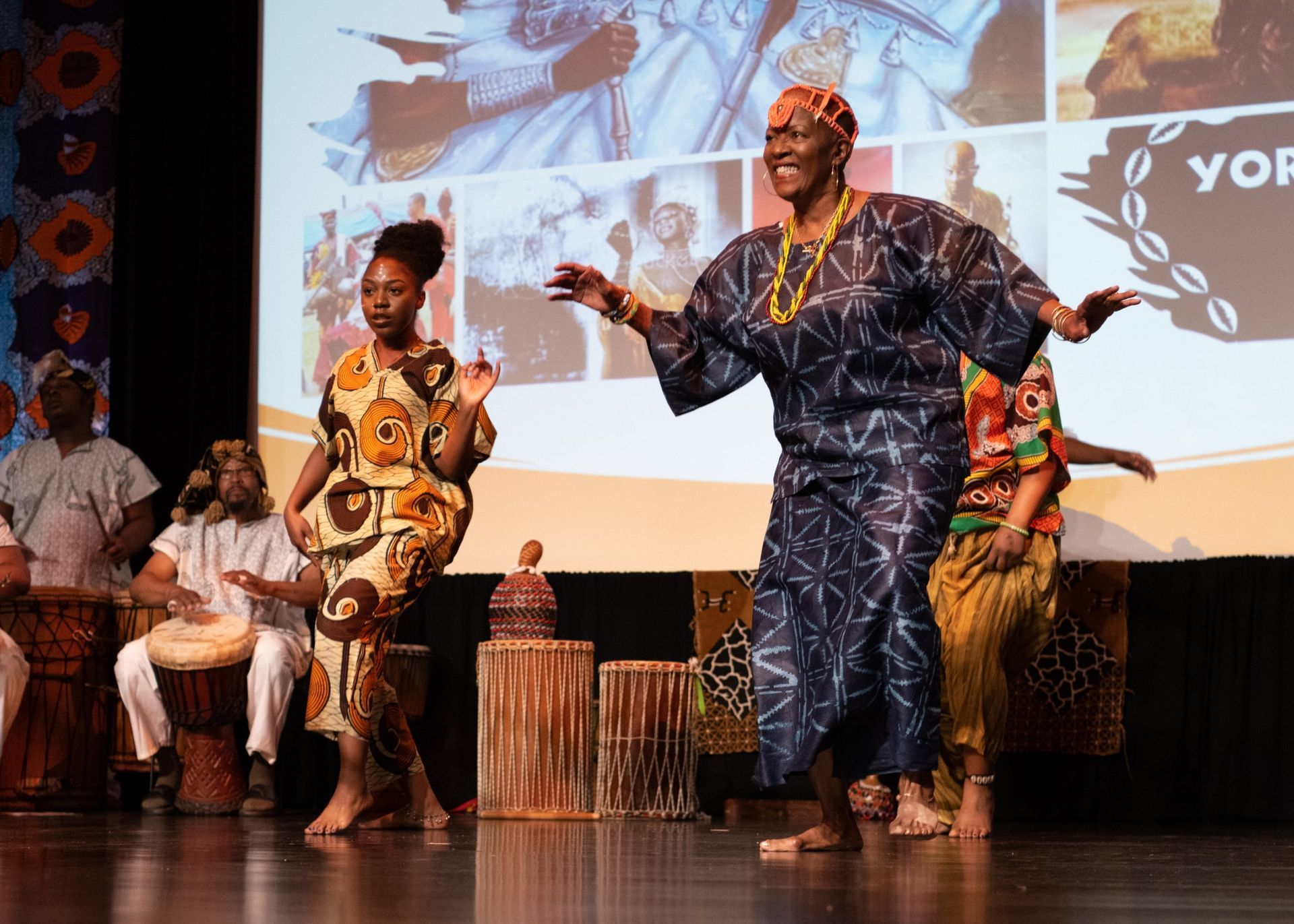 People in African attire dance on a stage. Drummers play. A screen with text and images is behind them.