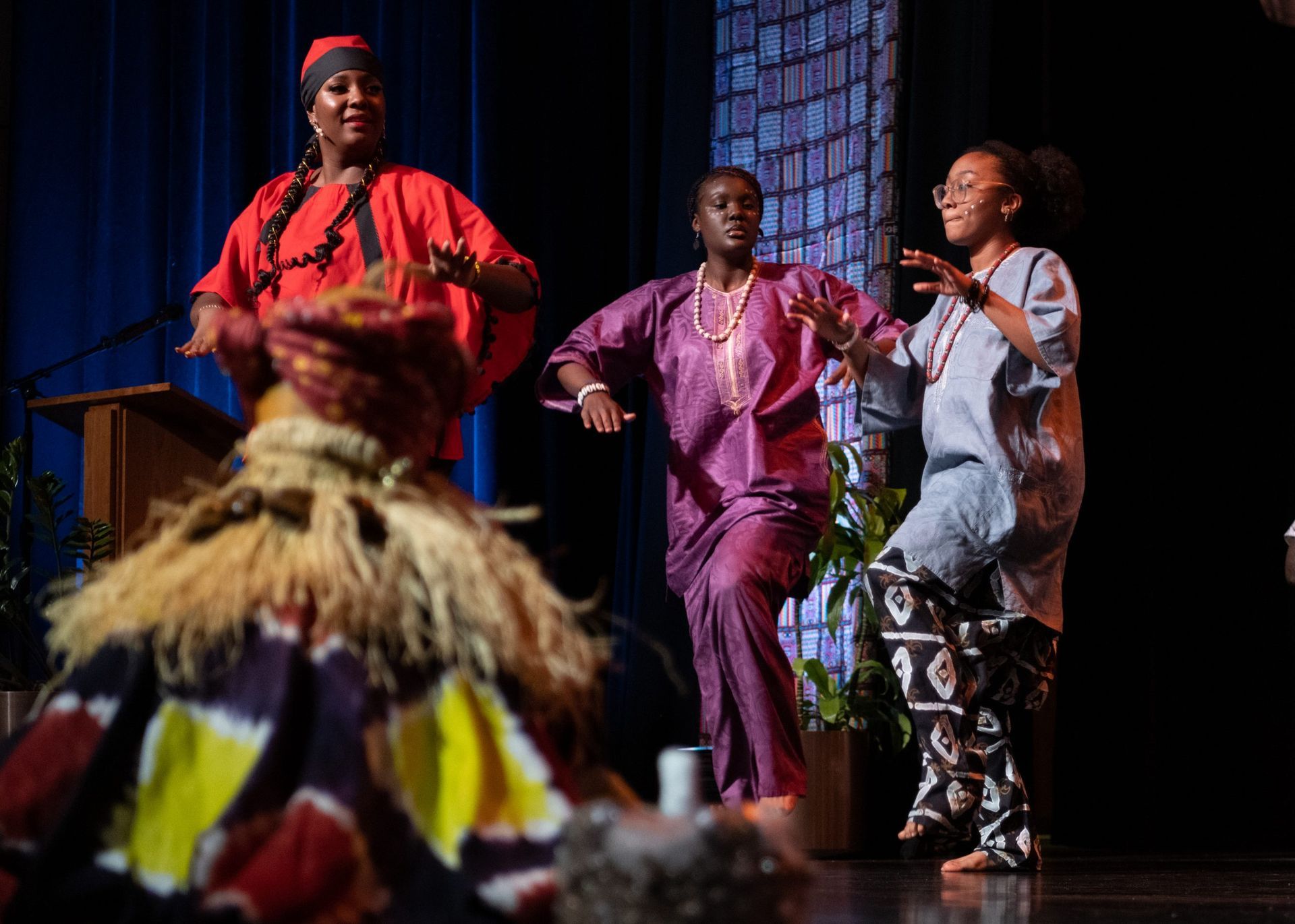 Three women in colorful African attire dance on stage, behind a decorative mask.