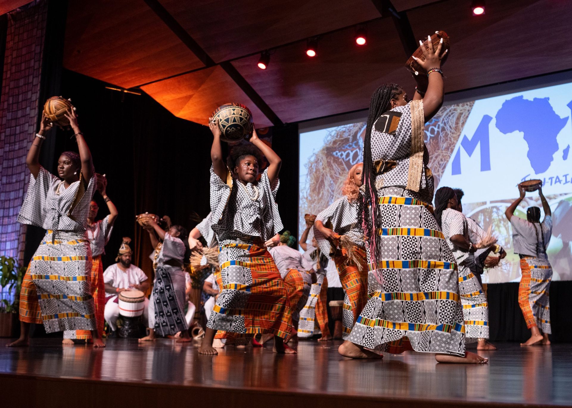 Dancers in colorful African attire on stage with props, raising arms. Backdrop with a map of Africa.