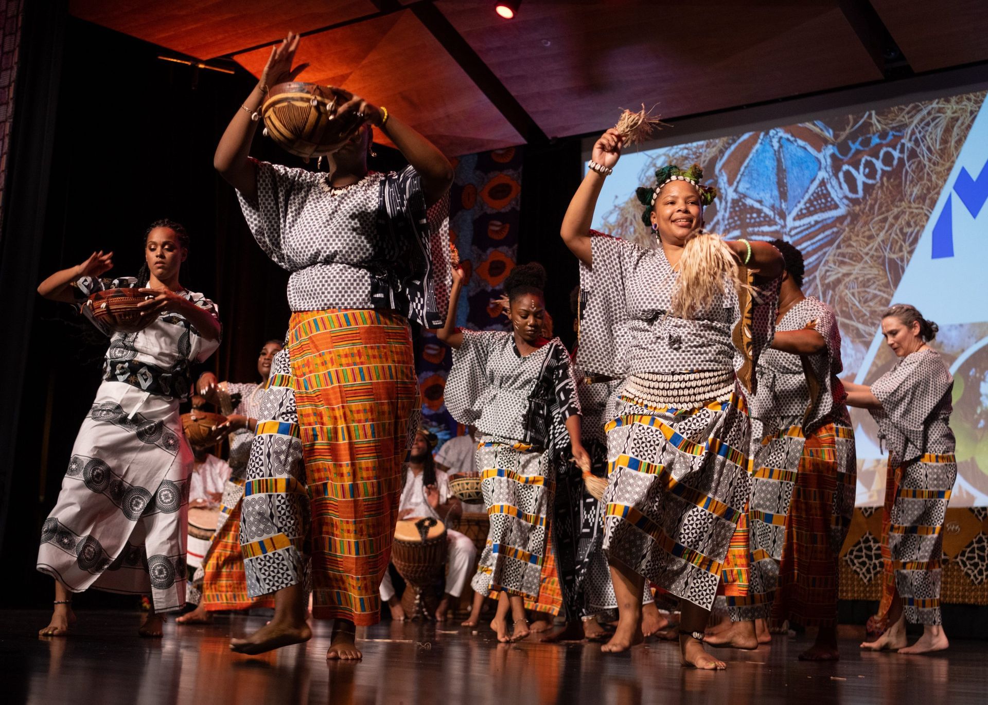 People in African attire dance on a stage with a colorful backdrop; drums are visible.