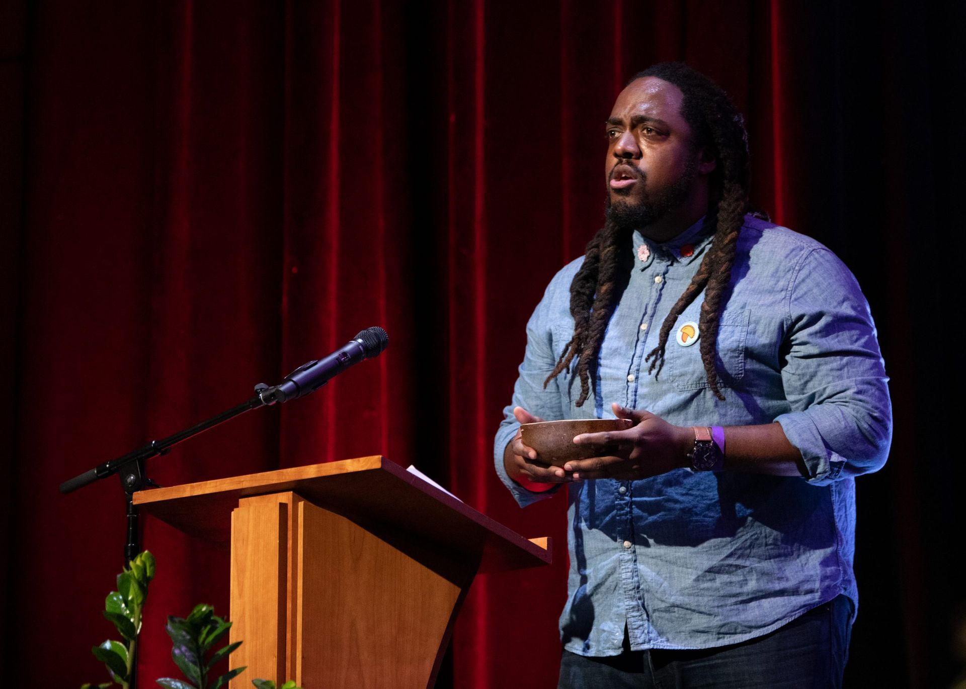 Man with dreadlocks speaking at a podium with a microphone, in front of a red curtain.