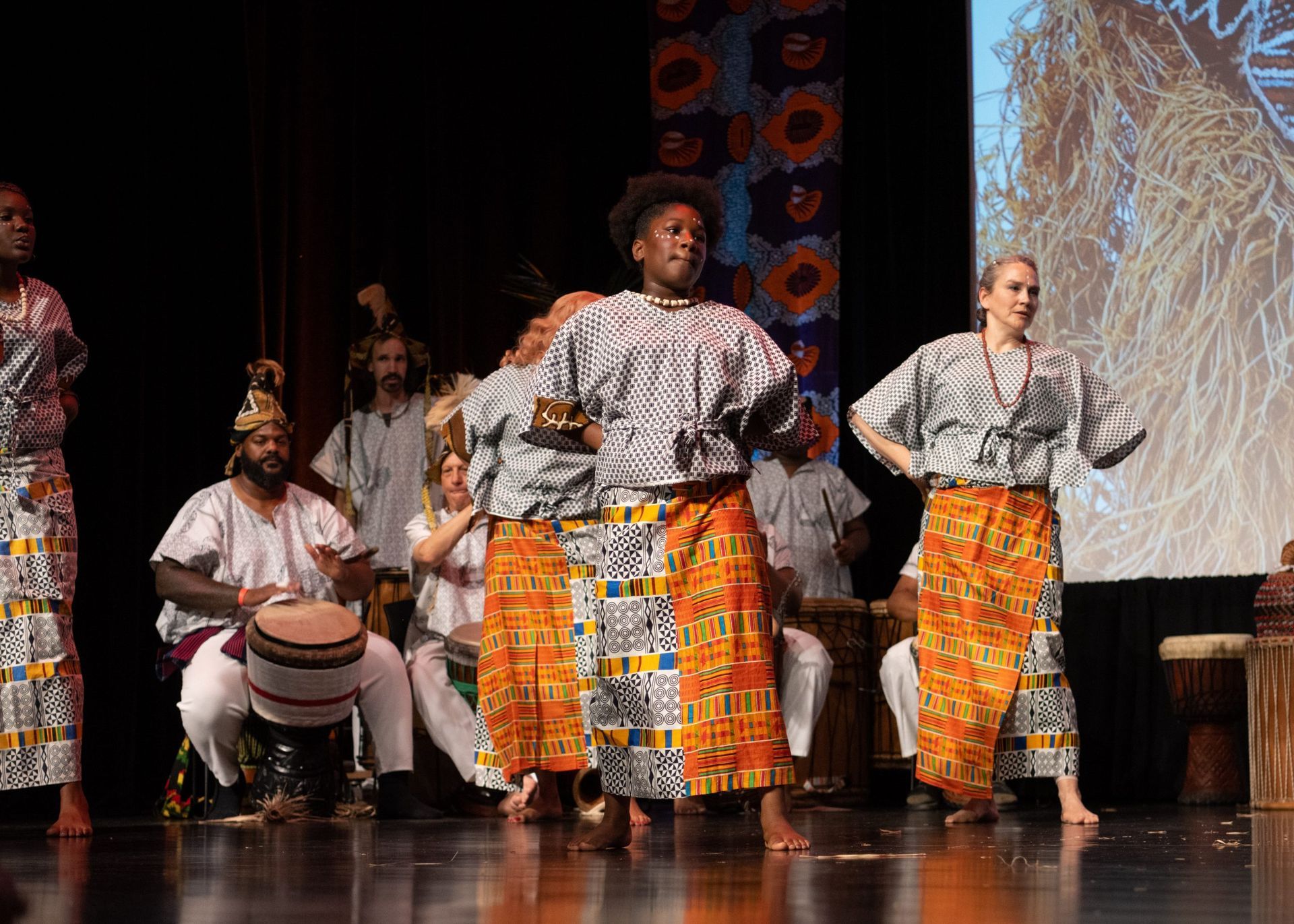 African dancers in traditional clothing on stage, musicians play drums in background.