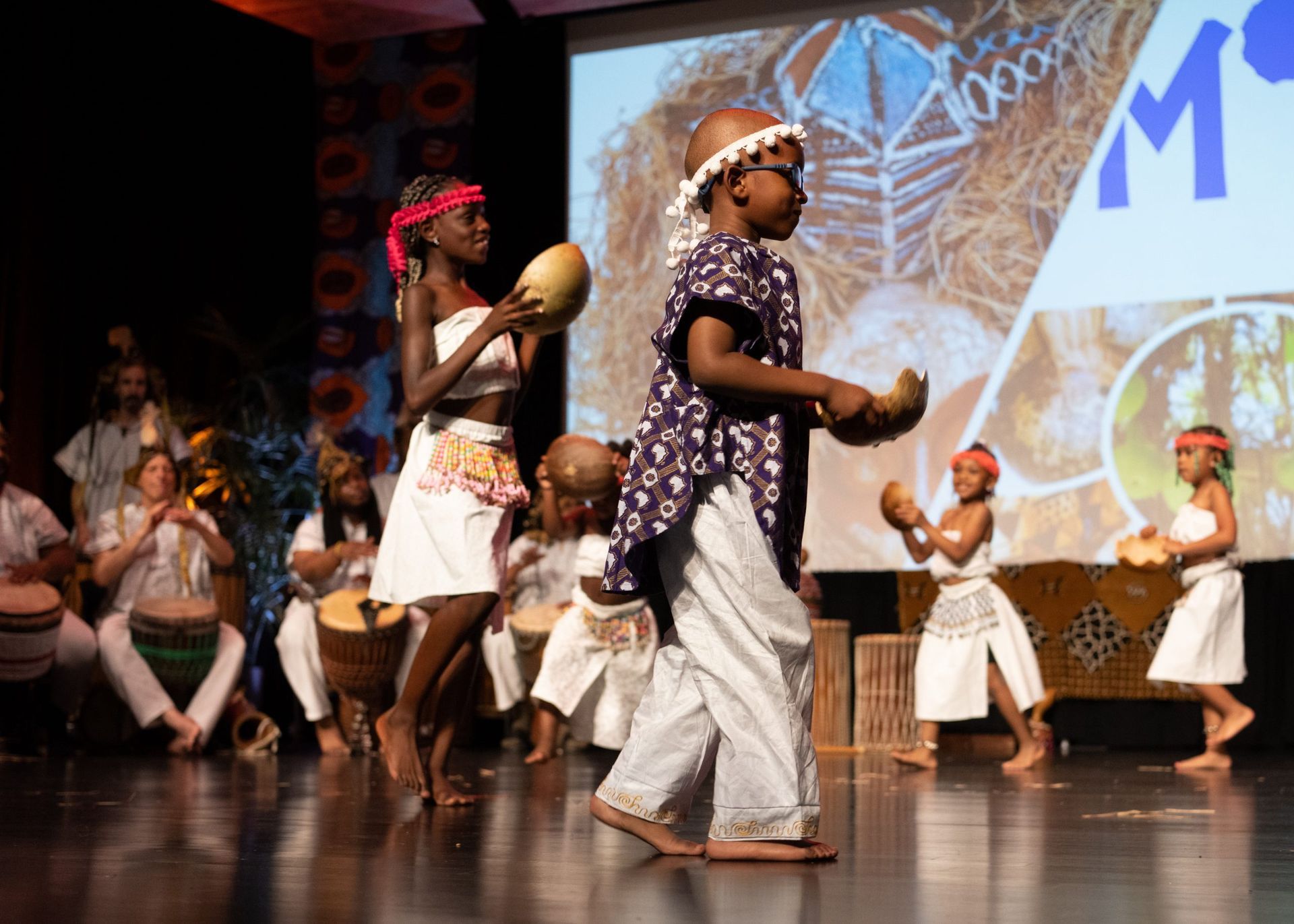 Children in traditional African outfits perform on stage. Some play instruments, others dance. Stage with projection.