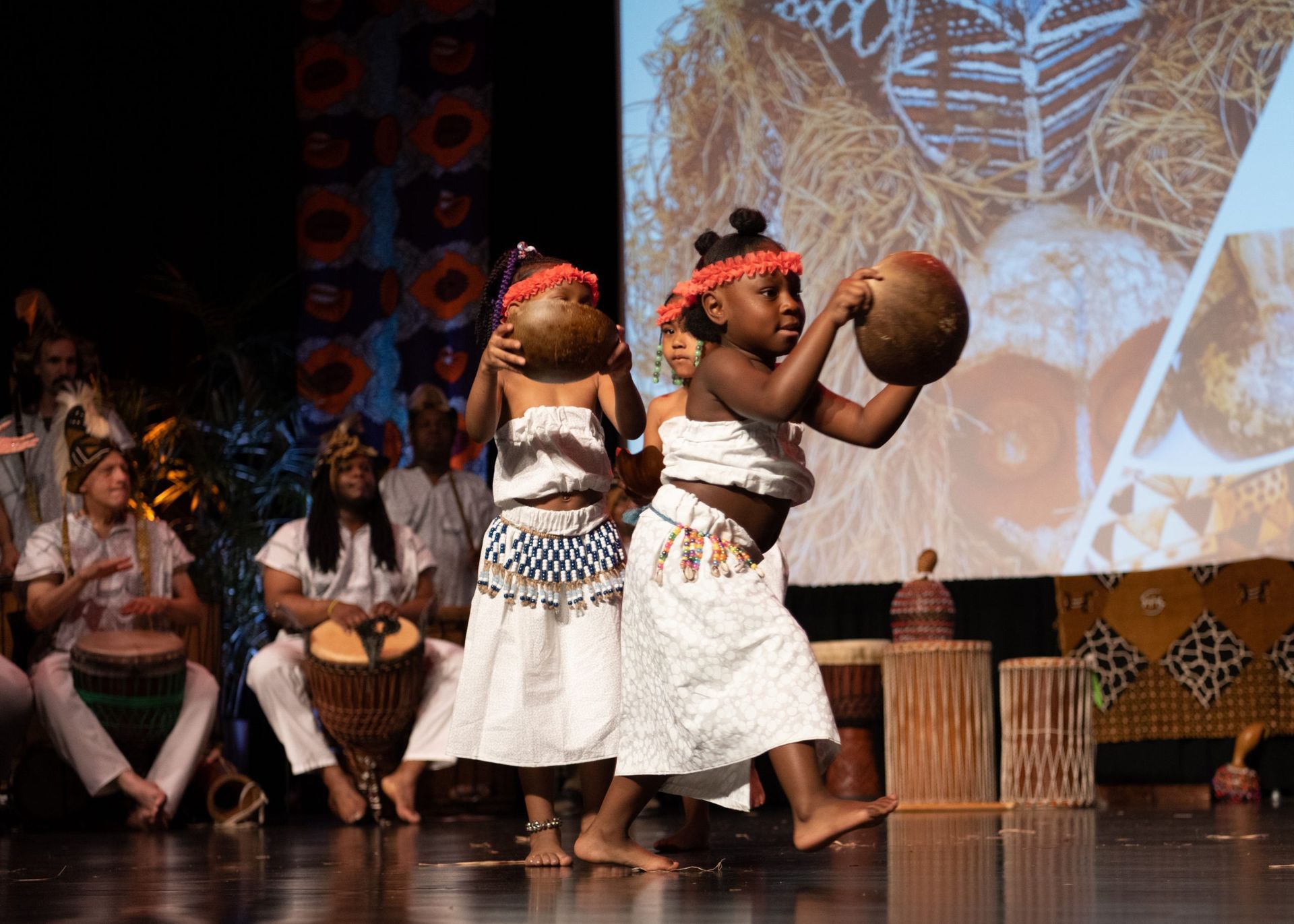 Two young girls dancing with gourds on stage, with musicians and cultural backdrop.