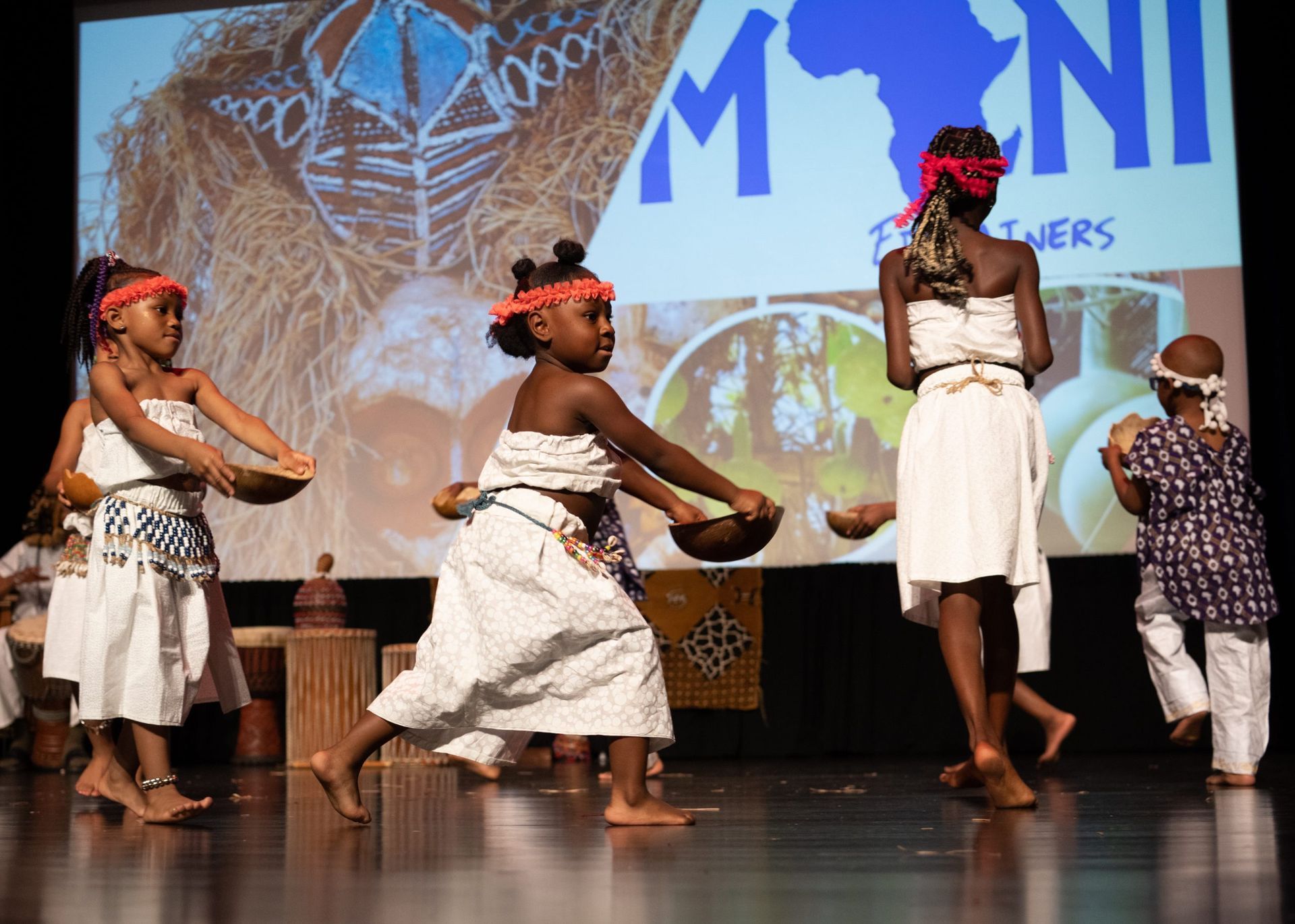 Children in white outfits dance on stage, holding bowls, with African-themed backdrop and drums.