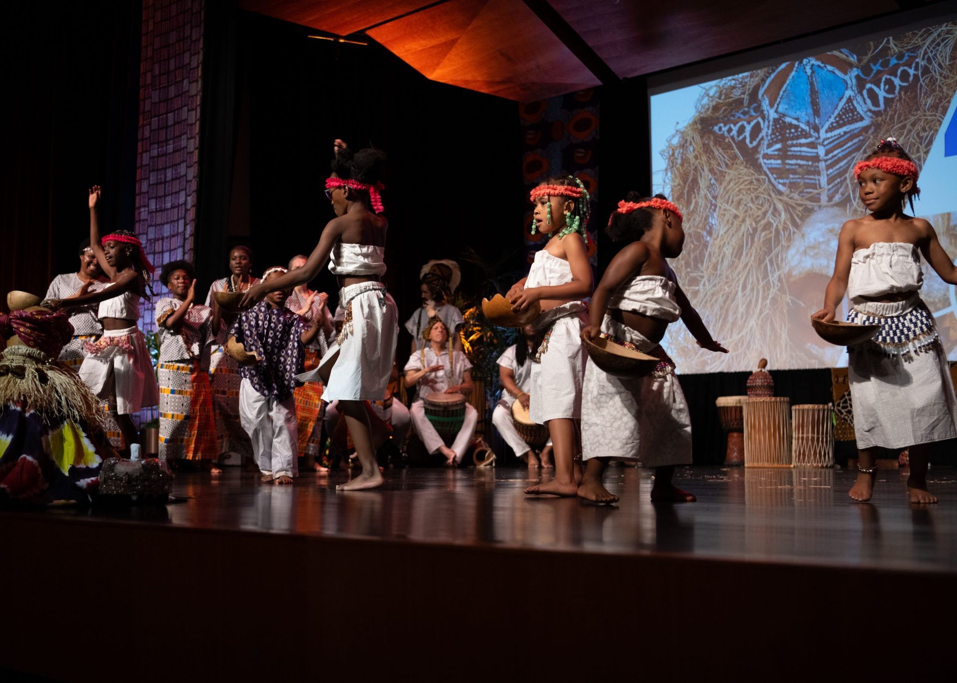 Children in white and colorful costumes perform a cultural dance on stage, lit by warm lights.