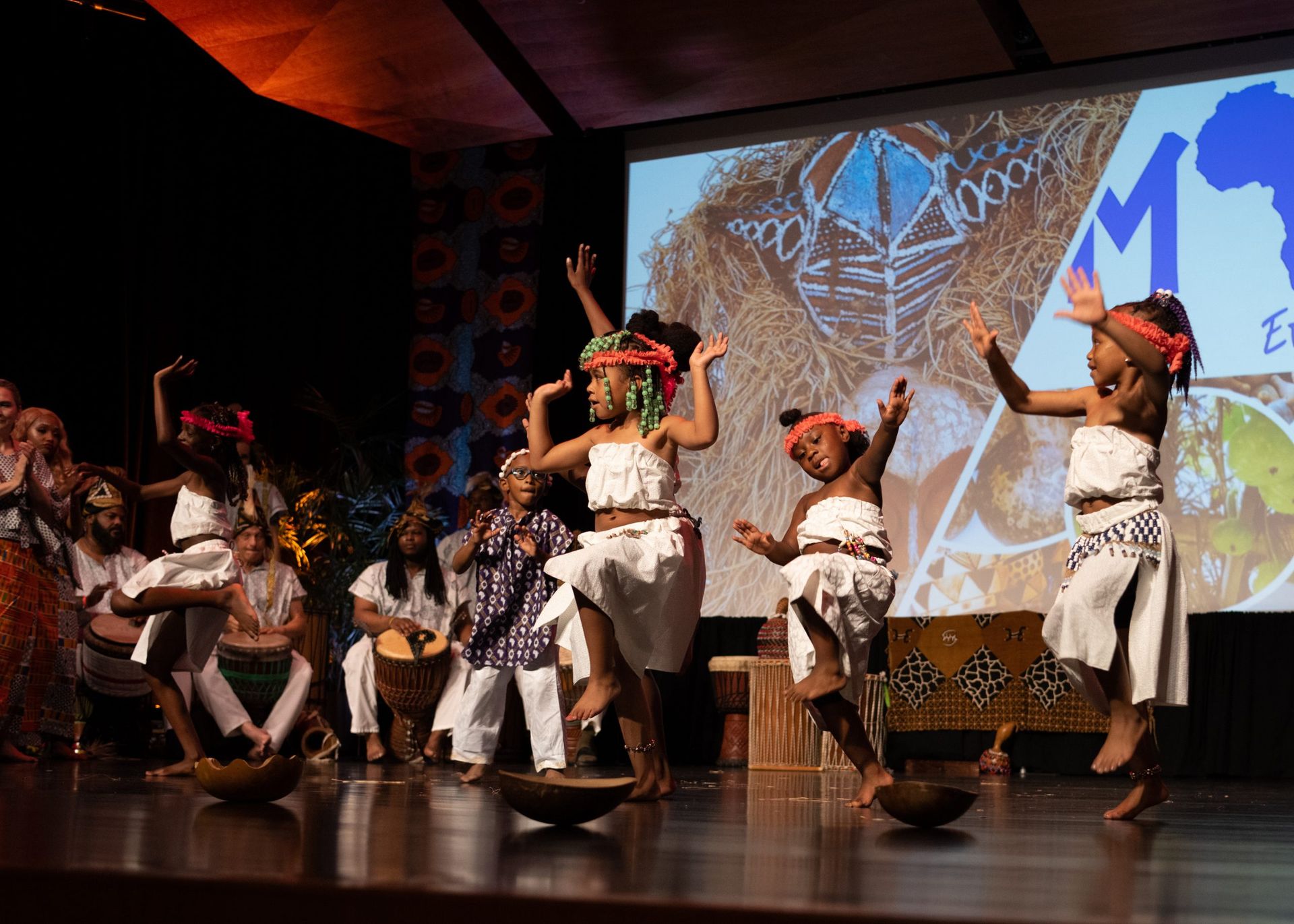 African dancers perform on stage with props and a backdrop.