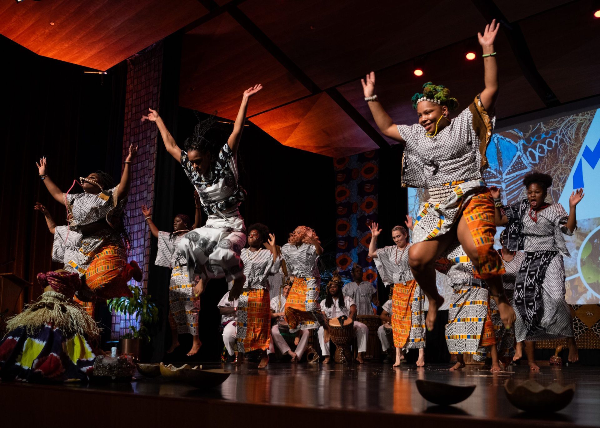 African dancers jump on stage, wearing colorful patterned clothes, under warm lights.