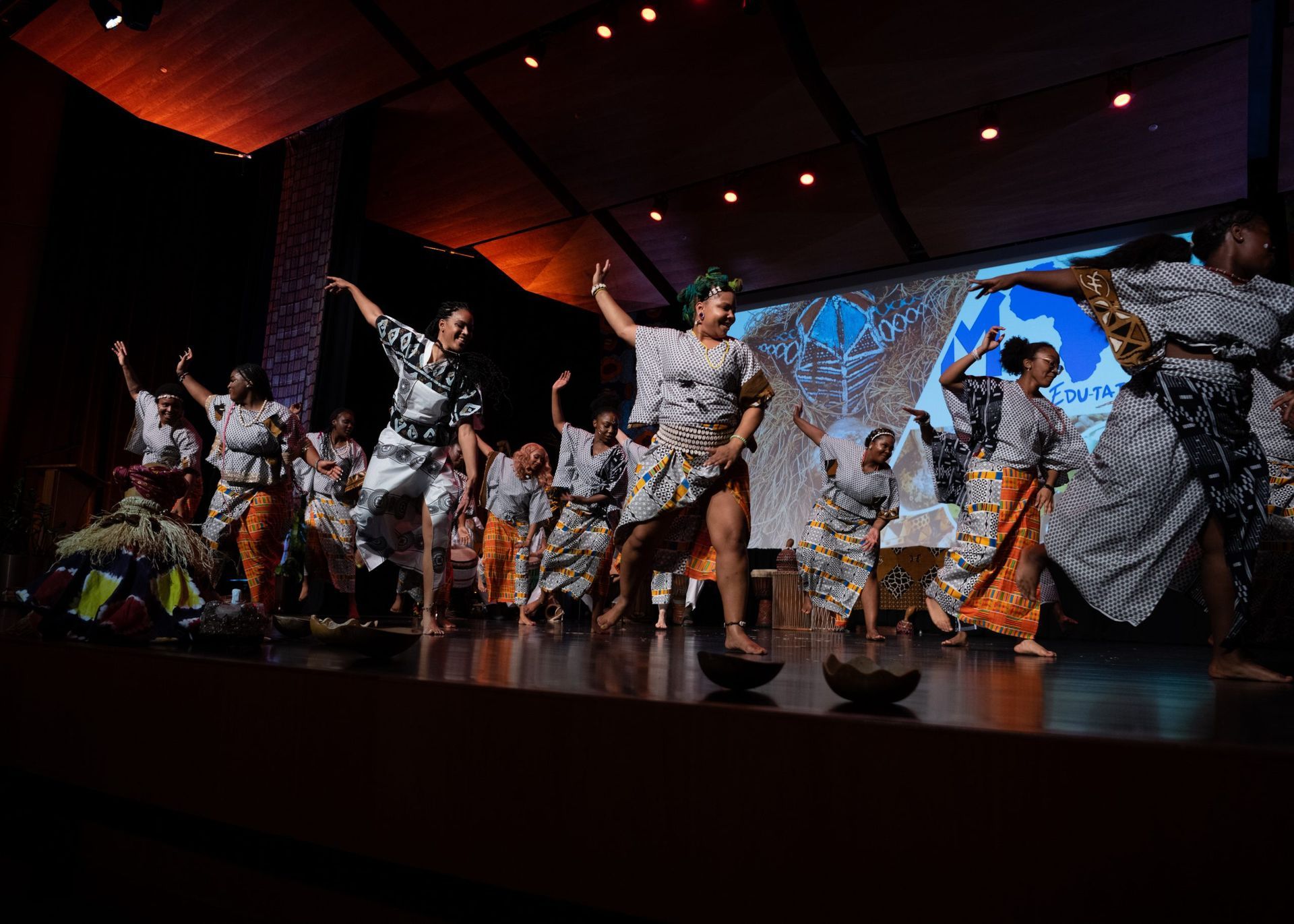 People in traditional outfits dance on a stage, arms raised. A light blue background.