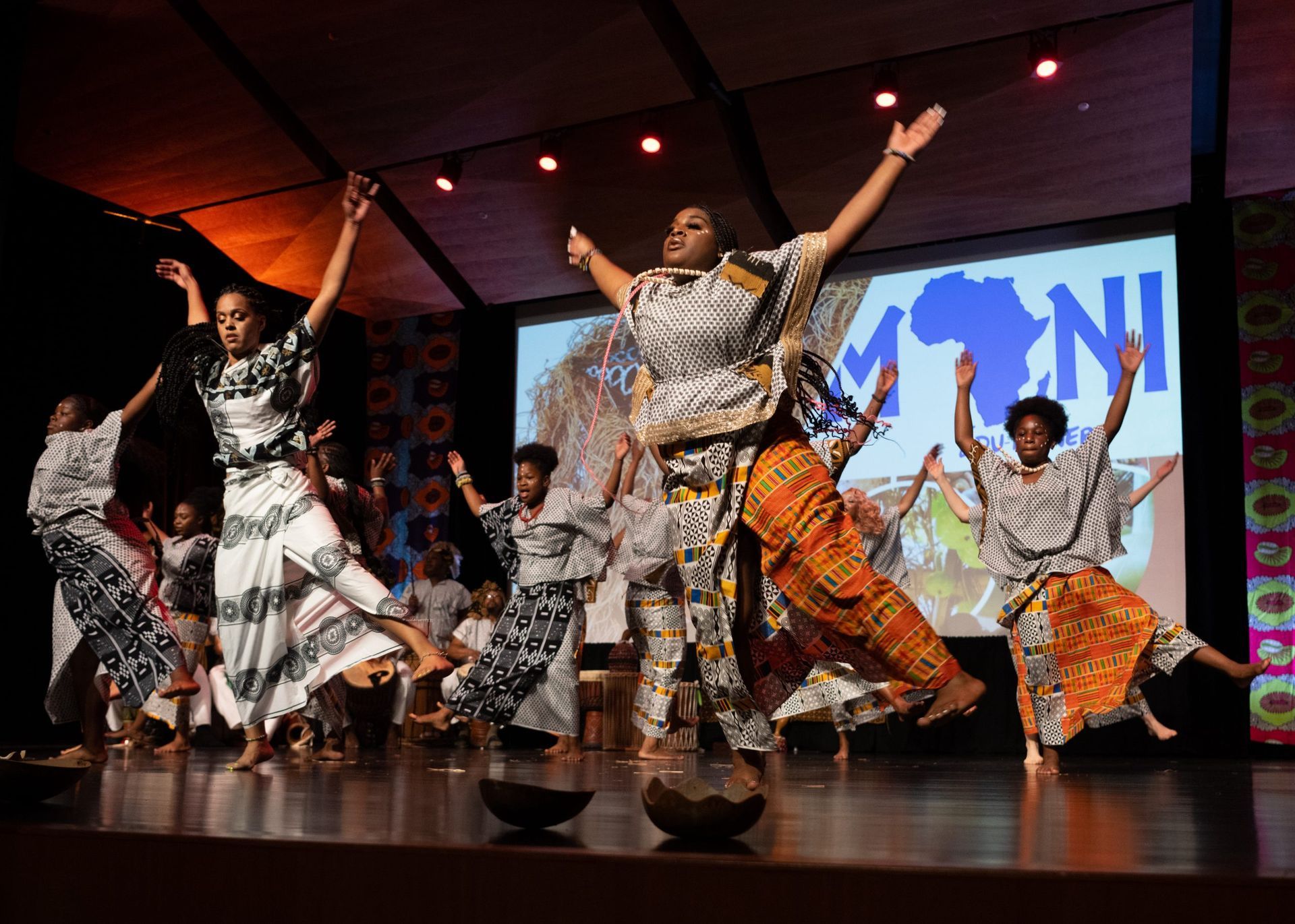 Group of people dancing on stage, arms raised. Wearing patterned outfits, in front of a screen.