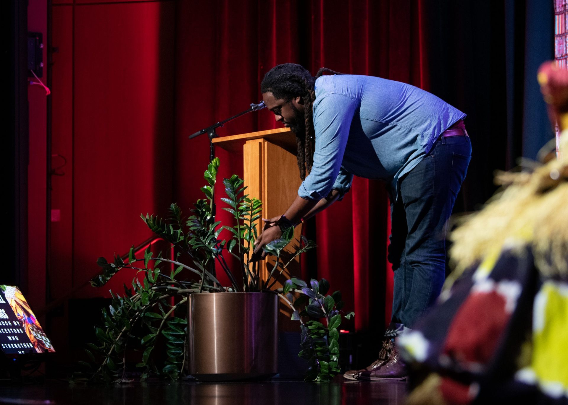 Man tending a plant onstage near a podium and red curtain.