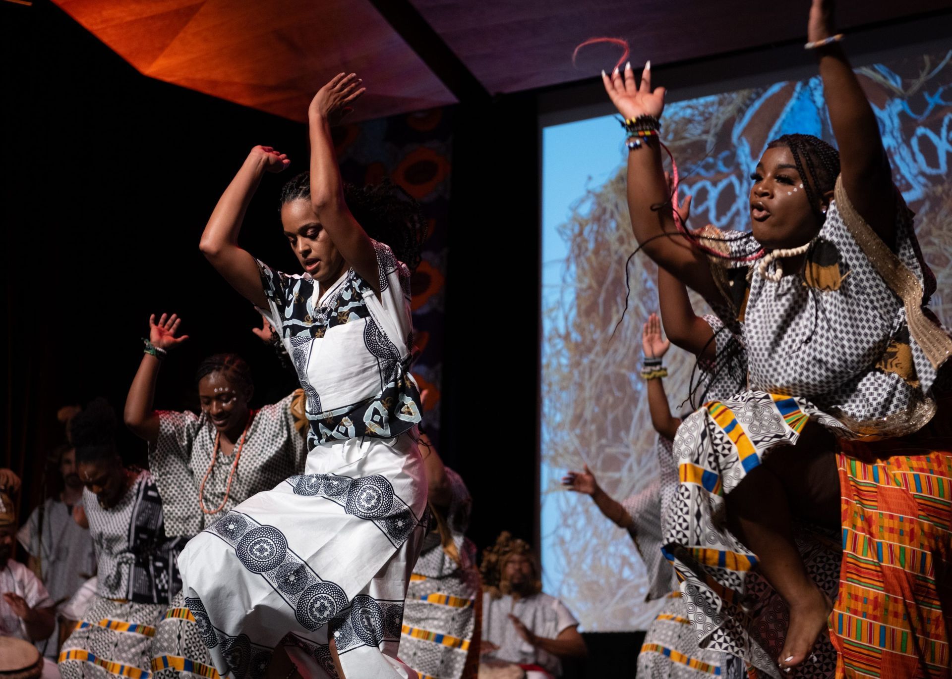 Dancers in patterned clothing jump with arms raised. Stage with projection screen and colorful decorations.
