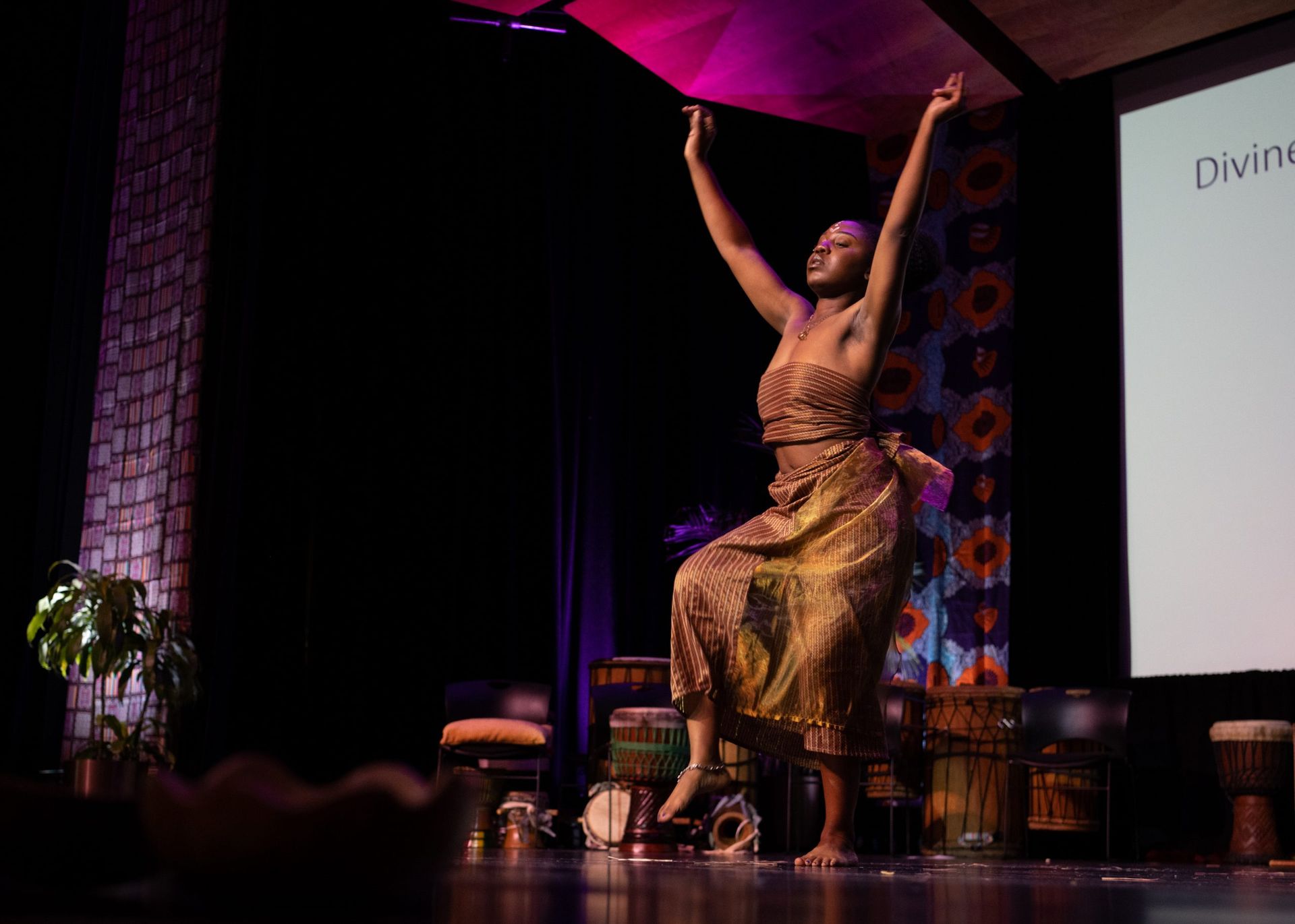 Woman in patterned dress dances onstage with arms raised; drums and screen in background.