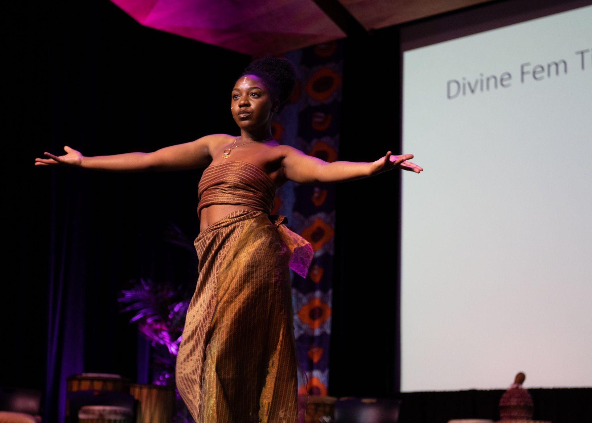 Woman in brown outfit dances on stage, arms outstretched. A screen displays 