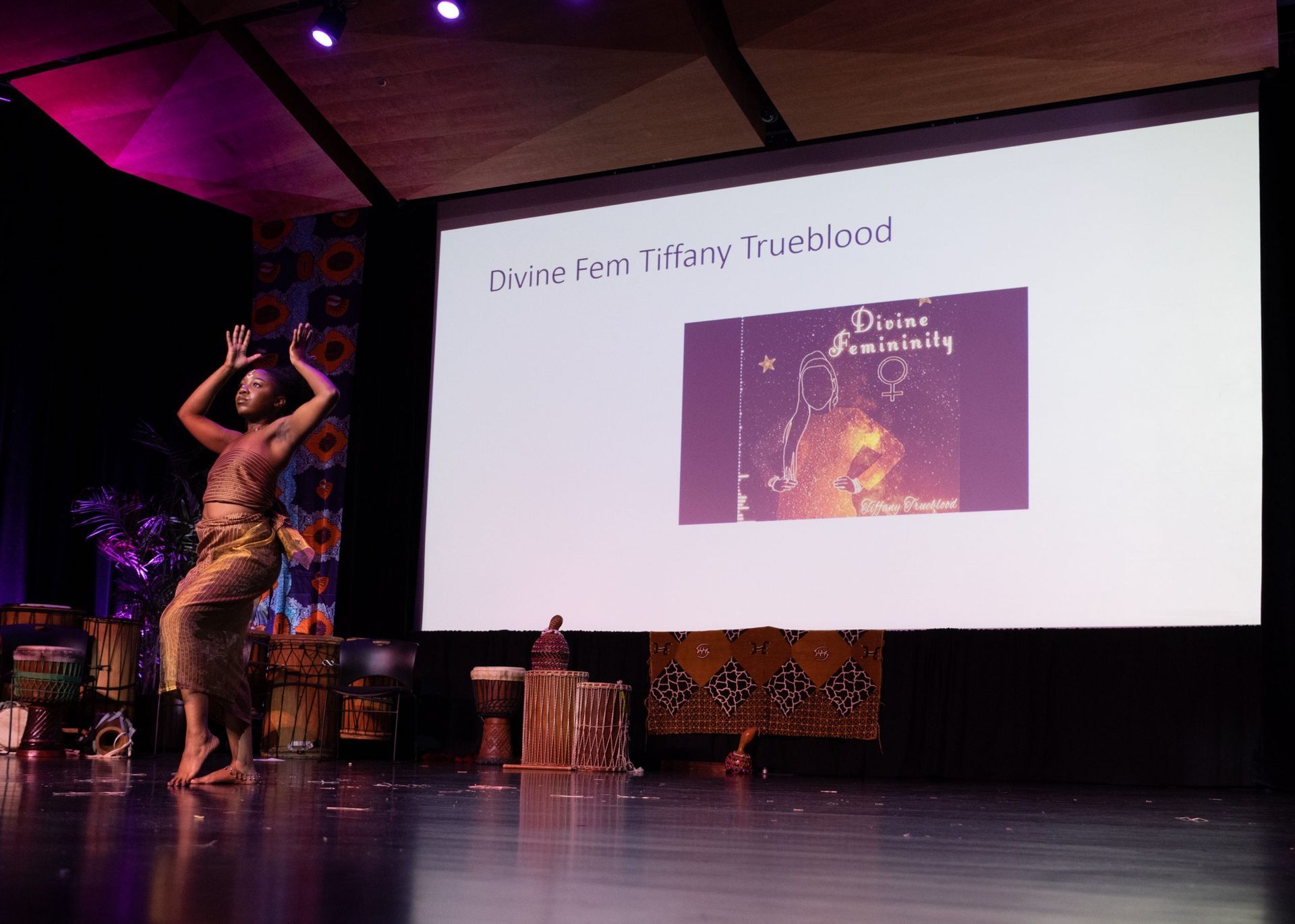 Dancer on stage performing in front of a screen displaying a title and image. Drums and decor flank the stage.
