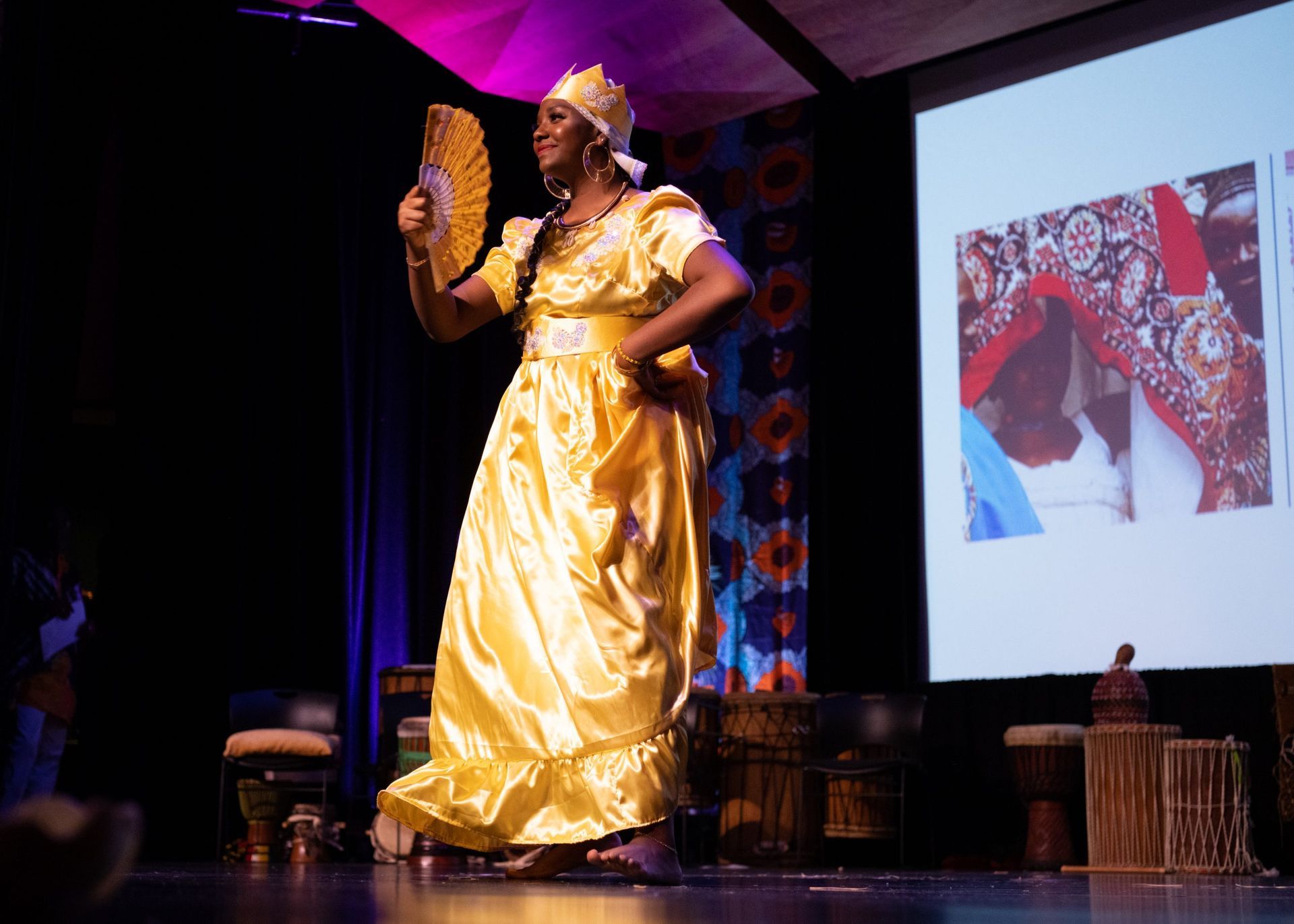 Woman in golden dress, holding fan, on stage with slideshow.